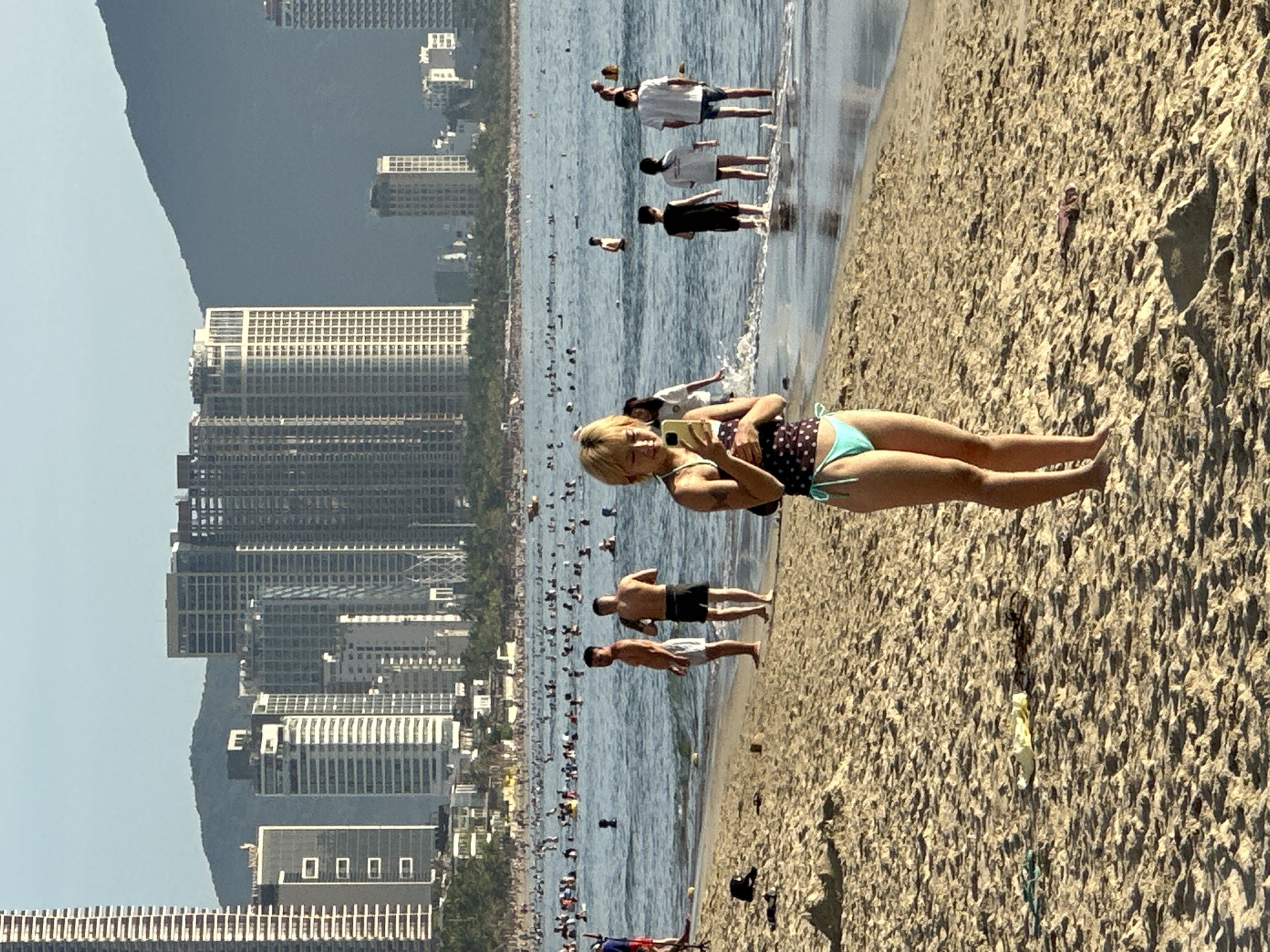 A crowd of people are enjoying a sunny day at a beach in Đà Nẵng, Vietnam. The shoreline is lined with sand, and in the distance, several tall buildings and mountains can be seen. In the foreground, a woman with blonde hair is standing on the sand, looking at her phone. She is wearing a polka-dot bikini with turquoise bottoms. Behind her, other beachgoers are scattered on the sand and in the water. Some are swimming or wading, while others are standing and talking. The water appears calm, with small waves gently rolling onto the shore. The sky is clear and blue, indicating good weather.
