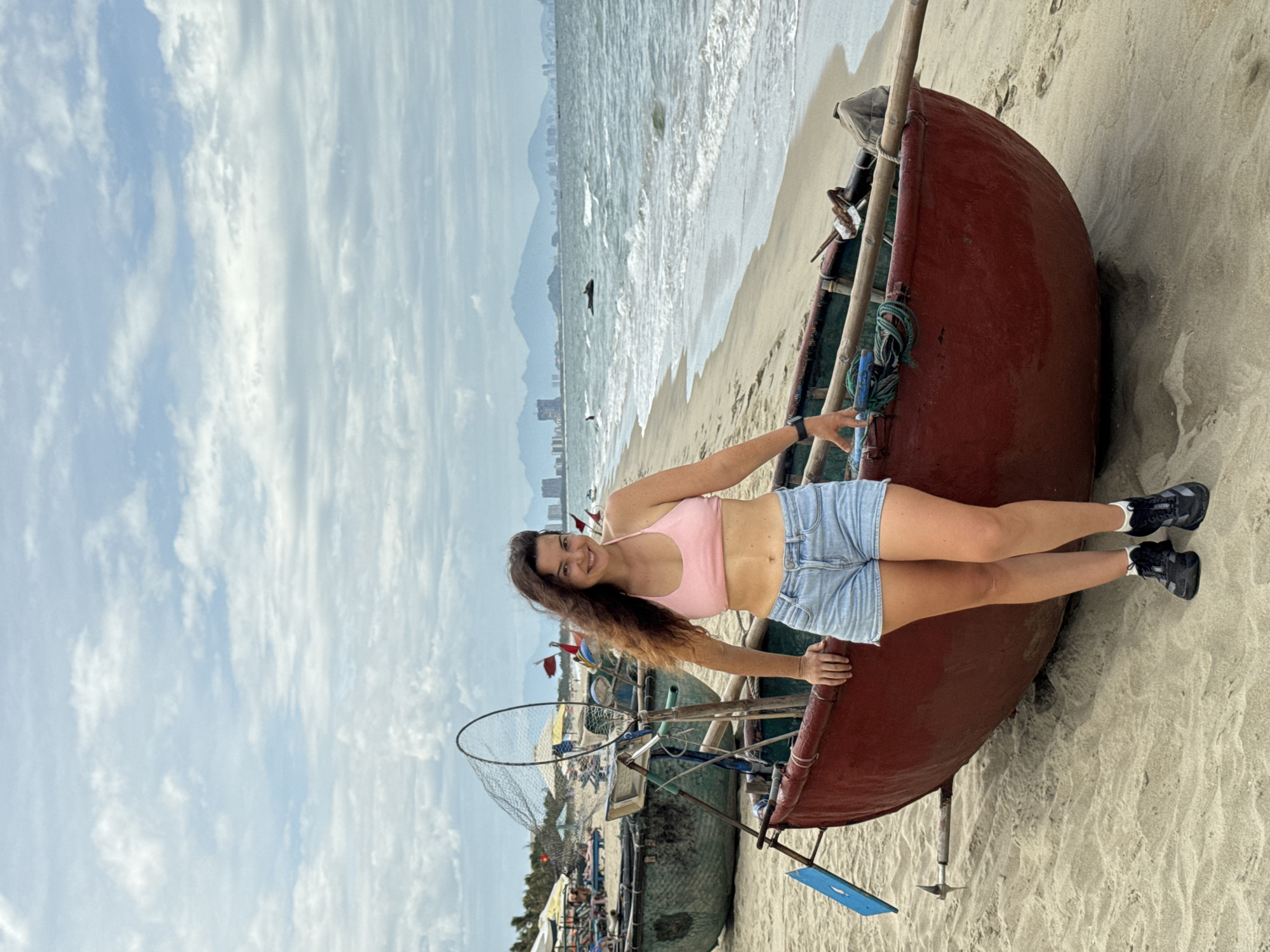 A woman stands on a sandy beach in Hoi An, Vietnam, leaning against a large, red, bowl-shaped boat. She is wearing a pink bikini top and light blue denim shorts, with black sneakers. Her dark, wavy hair is blowing in the wind. Behind her, more boats, fishing nets, and beach umbrellas are visible. In the distance, the ocean meets a hazy skyline with mountains. The sky is overcast with scattered clouds.
