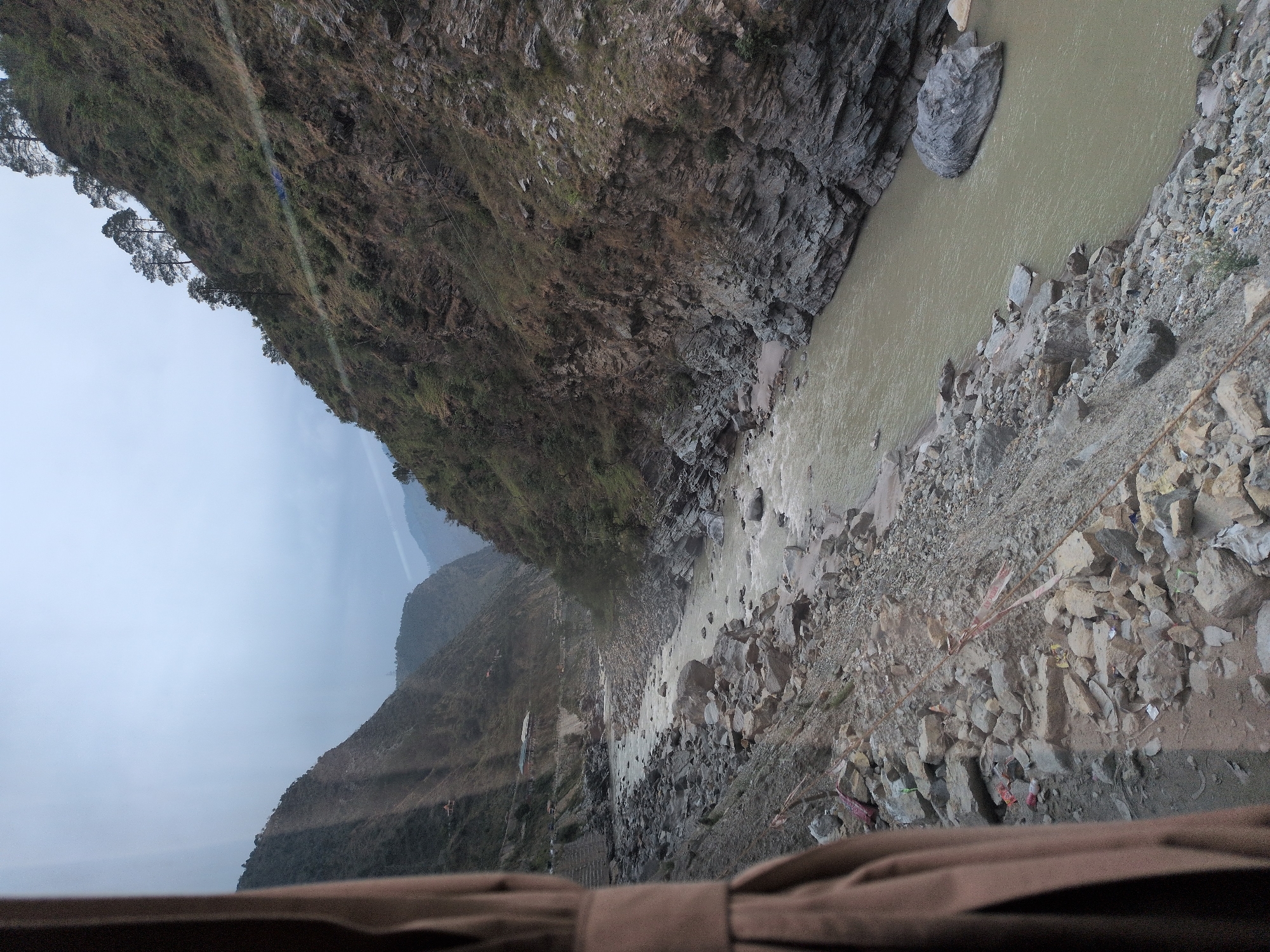 The image shows a murky river flowing through a rocky gorge in Gurahan, India. The riverbanks are steep and covered in rocks and sparse vegetation. Trees grow on the slopes of the gorge, and some buildings are visible in the distance along the river. The sky is overcast, giving the scene a somber atmosphere. A brown curtain is visible in the left foreground, suggesting the photo was taken from inside a vehicle.