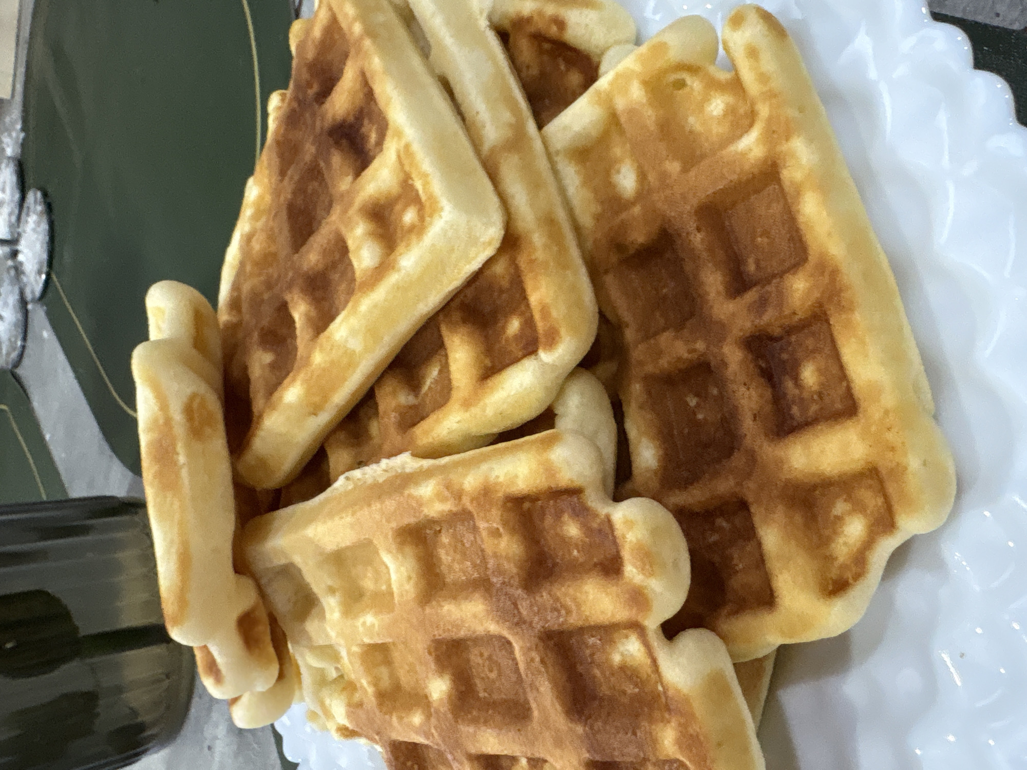 A close-up view depicts a stack of approximately ten rectangular waffles piled on a white, scalloped-edge plate. The waffles vary in color from light golden-yellow to darker golden-brown, displaying a distinct grid pattern on their surfaces. The background is blurred, showing a dark green surface, possibly a placemat or tablecloth, on which a dark, fluted container is partially visible on the left. Faint, glinting reflections, indicative of glassware, are also present on the green surface to the right. The scene is consistent with an indoor, domestic setting within the city of Chaikovsky, Russia. No individuals are present or engaged in any activity.