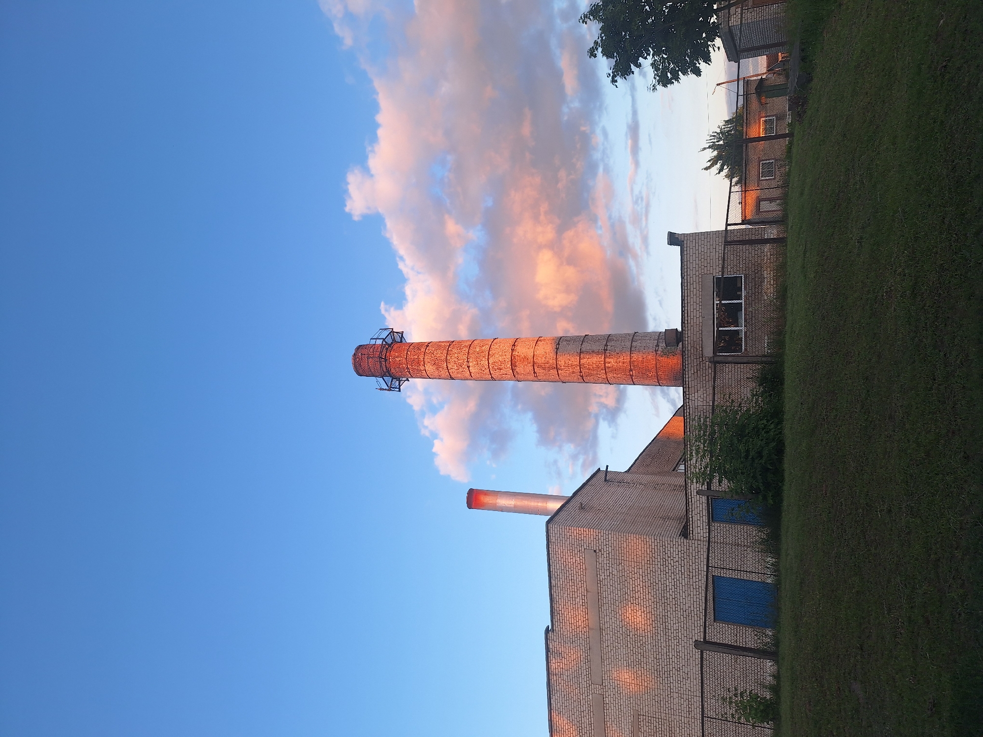 This is a photograph of an industrial facility, likely a former or current factory, located in the Капыльскі сельскі Савет, Belarus. The sky is a vibrant blue with scattered clouds illuminated by the setting sun, casting warm hues of pink and orange onto the scene.
In the foreground, a grassy slope leads up to a chain-link fence. Behind the fence, a long, light-colored brick building dominates the left side of the frame, with a smaller, equally brick building to its right. Two blue metal doors are visible on the building's exterior.
The most prominent feature of the facility is a tall, cylindrical brick chimney rising from behind the main building, illuminated by the warm light of the setting sun. To the left of this main chimney, a thinner, silver-colored chimney extends upward from the roof of the brick building. A few trees are visible in the background, silhouetted against the sky.
The overall impression is one of an aging industrial landscape bathed in the soft light of dusk, suggesting a quiet or abandoned state. There are no people or animals visible in the image.
