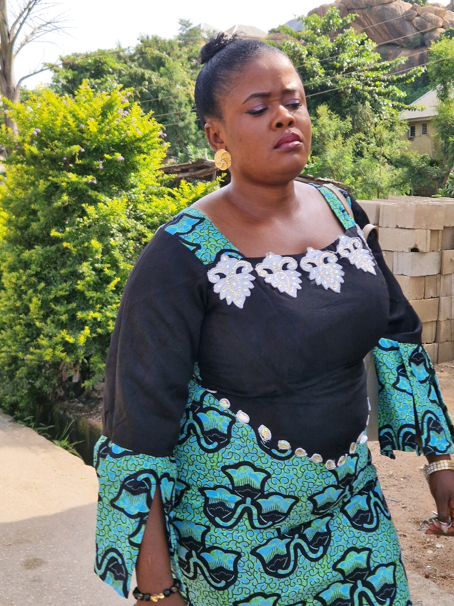 A full shot photo shows a woman standing on a path in Kabong, Nigeria. She is wearing a black dress with light blue and green patterns. The woman has dark skin, her hair is pulled back in a bun, and she is wearing gold earrings. The background shows a leafy green bush, a large rock formation, and some buildings. In the foreground, there are stacks of grey cinder blocks. The lighting suggests it is daytime.