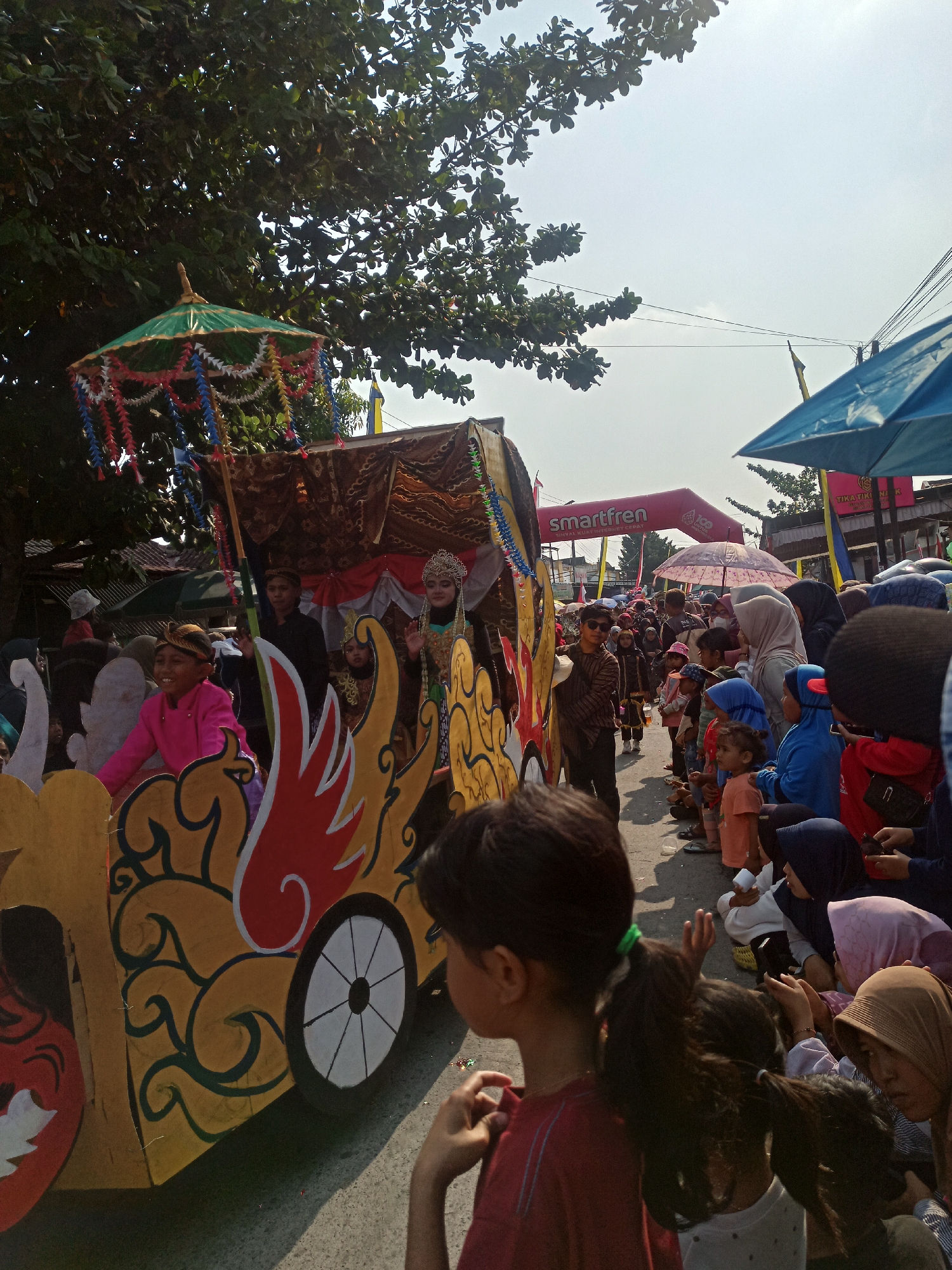 An outdoor parade is taking place on a paved street in Saren, Indonesia. A decorated float resembling a chariot, adorned with gold, red, and green motifs, is the central element. Several individuals, including children and adults, are dressed in traditional attire and appear to be participating in the procession. A young boy in a bright pink garment is seated on the float and smiling. On the float, a person wearing a jeweled headdress and a gold and blue outfit is waving to the crowd. Spectators, including many children and women wearing headscarves, are gathered along the roadside, some sitting on the ground and others standing, watching the parade. Umbrellas, some blue and some pink, are visible, providing shade. A banner advertising "Smartfren" is also present. The sky is bright and clear, indicating a sunny day. Trees with dense foliage line the background, and buildings are visible in the distance. The overall atmosphere appears festive and celebratory.