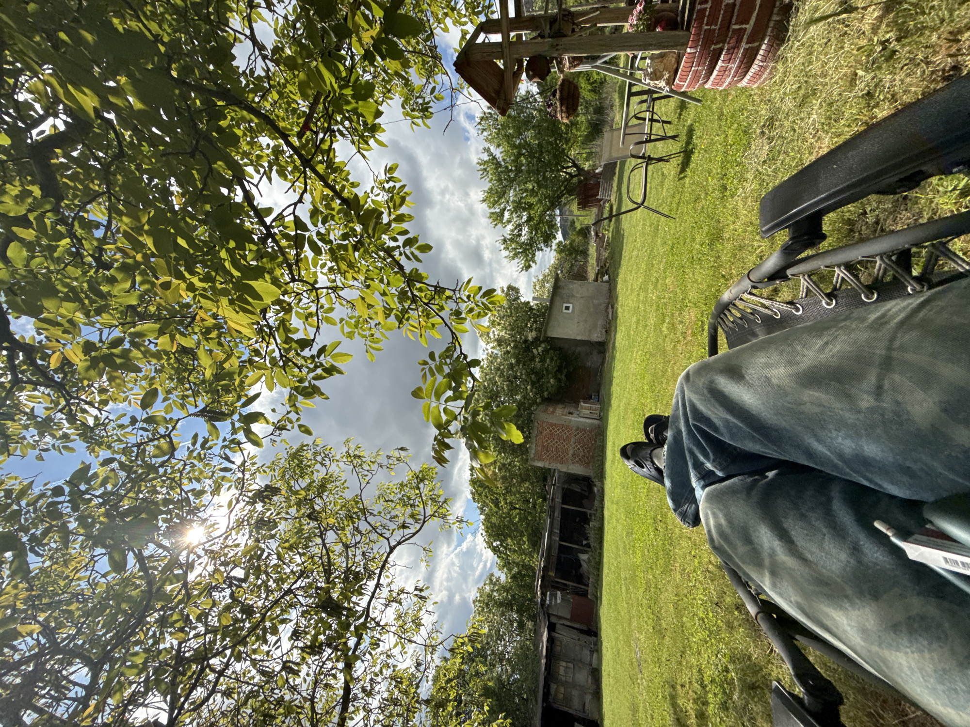 The image is a low-angle, outdoor shot taken from the perspective of someone sitting in a reclining chair. The person's legs, clad in faded denim jeans and black footwear, are partially visible in the foreground on the right side of the frame. The background reveals a grassy lawn, trees with green leaves, and a partly cloudy sky with patches of blue. In the middle ground, there's a dilapidated shed or outbuilding. Further back, garden furniture and decorative items are visible, possibly a swing set, chairs, and a brick structure. The scene appears to be a backyard or garden area, likely in Sliven, Bulgaria. The image has been rotated and should be re-oriented for better viewing.
