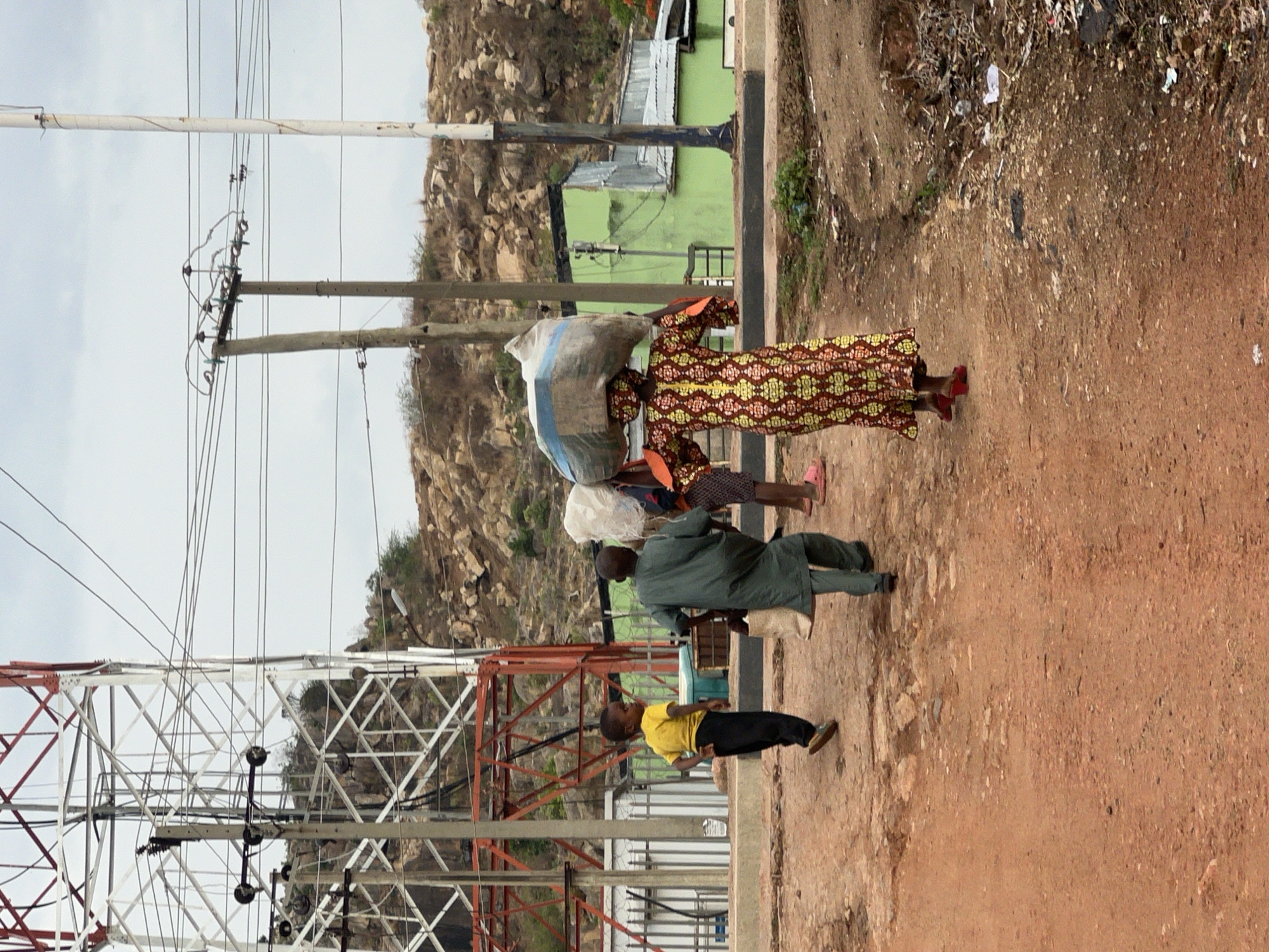 This media file captures a daytime scene in Gura Top, Nigeria, showing several individuals walking along a reddish-brown dirt path. The setting appears to be a semi-rural area characterized by rocky, sparsely vegetated hills in the background.
In the foreground and midground, a group of people is visible. Closest to the viewer, on the left, is a child wearing a yellow t-shirt and dark pants, walking to the right. Behind them, a man in a dark green traditional garment also walks right, while further back, a woman in a brightly patterned dress (featuring red, yellow, and brown/green motifs) balances a large, light-colored sack or bundle on her head. She wears red footwear. Another person, partially visible behind her, also carries a white bundle on their head, suggesting the transport of goods.
The scene is rich with infrastructure and natural elements. On the far left, a large, red and white metal lattice tower, possibly a communication or electricity transmission tower, dominates the lower left portion of the image. Multiple wooden utility poles run vertically through the center of the frame, laden with a dense network of electrical or communication wires. In the background, beyond the utility poles, glimpses of simple buildings are seen, including one with a distinctive light green wall and a metallic roof. The sky appears overcast, providing diffused light and casting no harsh shadows. The ground along the path shows signs of minor debris or litter. No visible text is discernible in the image.