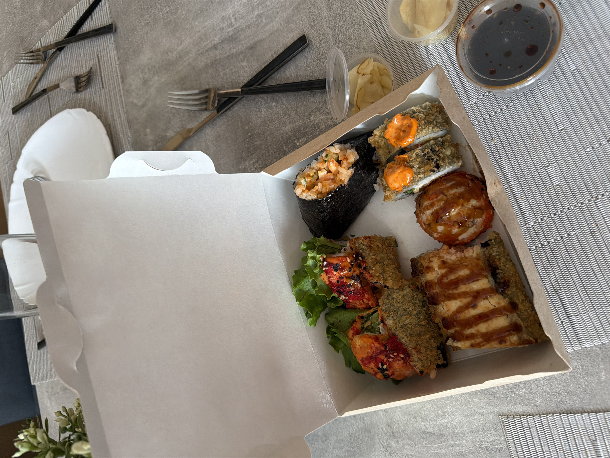 An overhead shot shows a takeout sushi box opened on a gray, textured placemat. Inside the box, there are several sushi rolls and one sushi hand roll. One roll is topped with a red spicy sauce and sesame seeds. Another is topped with bright orange sauce and appears to be fried. A hand roll is visible on the right side of the box. There are also two small plastic containers, one filled with pickled ginger and another with soy sauce. In the background, there are two white plates and a glass of water on a gray table. There are also two sets of forks and knives, suggesting a meal for two. The scene appears to be indoors, possibly a dining area. There is no visual information that indicates this is in the city of Semiluki, Russia.