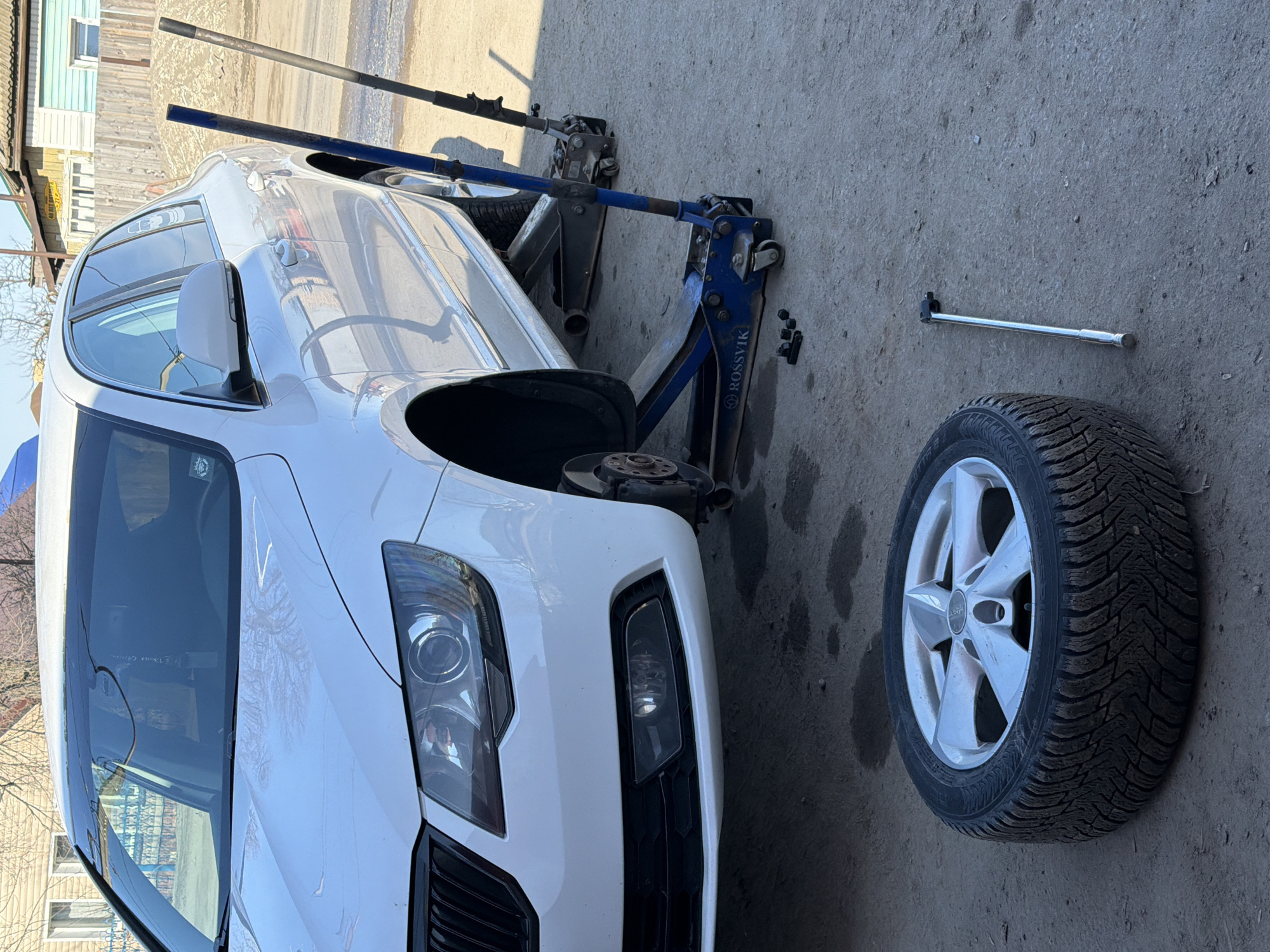 The image shows a white car undergoing a tire change outdoors. The front left wheel has been removed, and the car is raised on two jacks – one black and one blue – with a silver lug wrench lying on the ground near the removed tire. The tire, featuring a silver rim and winter tread, rests on the ground beside the car. A small building, possibly a house or shed, is visible in the background, suggesting a residential or semi-residential area. Based on the logo on the blue jack saying ROSSVIK, and the location in the question, the location of the photo is Chaikovsky, Russia. The scene suggests a do-it-yourself car maintenance activity.