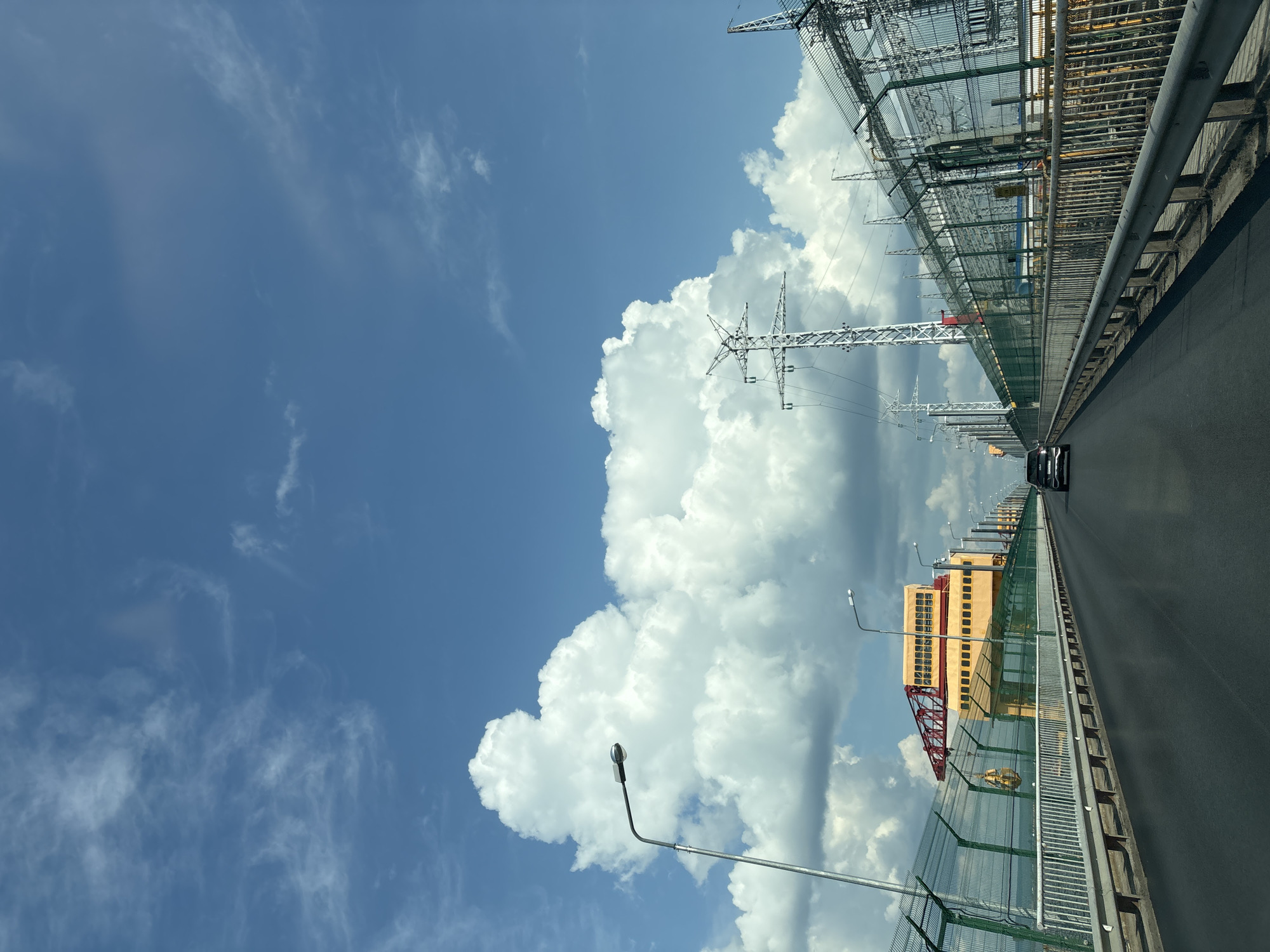 The image depicts a view from a vehicle driving on a bridge over a body of water. The bridge has a paved road with guardrails on both sides, separated by a metal fence topped with barbed wire. On the left side of the bridge, there are streetlights and a yellow industrial building with a red crane. On the right side, there are power lines and electrical substations, also enclosed by fencing. The sky is blue with scattered white clouds. The bridge is located in the city of Chaikovsky, Russia, likely over the Kama River, adjacent to the Votkinsk Dam and hydroelectric power station. A black car is visible ahead on the bridge. The scene appears to be an industrial area with significant infrastructure for power generation and transportation.