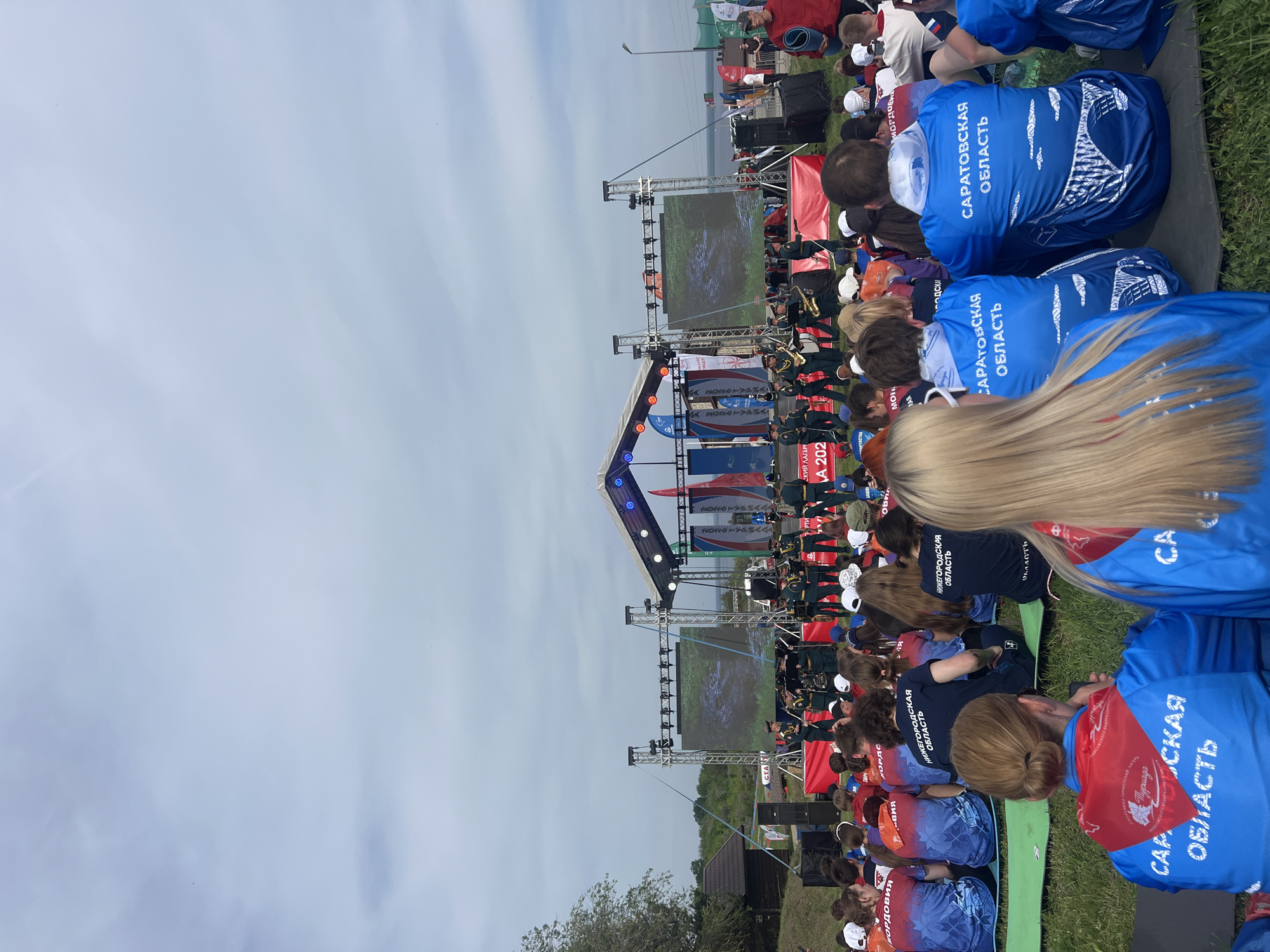 The image captures a view of a crowd of people sitting on the grass, observing an outdoor performance or presentation in Khvalynsk, Russia. The event appears to be well-attended. Many of the attendees are wearing blue shirts with the inscription "САРАТОВСКАЯ ОБЛАСТЬ" ("Saratov Oblast" in English). There's a stage with banners and screens displaying content, possibly related to the event. A band is performing on stage. There are tents on the upper left of the image, and a hut on the lower left. The sky is overcast.