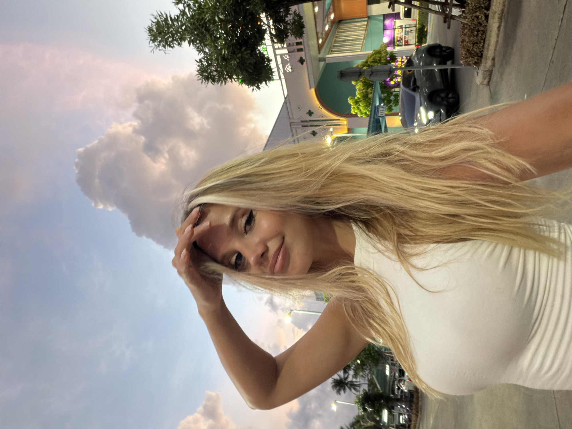 A young woman with long blonde hair is taking a selfie outdoors at dusk. She is wearing a white, form-fitting tank top. She has her right hand on her forehead and is looking at the camera with a slight smile. Her makeup includes eyeliner and lip gloss.
In the background, there is a sky with dramatic clouds, tinged with pink and orange from the setting sun. A modern building with green and white accents is visible, possibly a shopping center. To the right, there is a tree with lush green foliage. A dark-colored car is parked in the middle distance on a paved road. Palm trees are visible on the left side of the image. The scene appears to be in an urban or suburban area of Si Sunthon, Thailand.