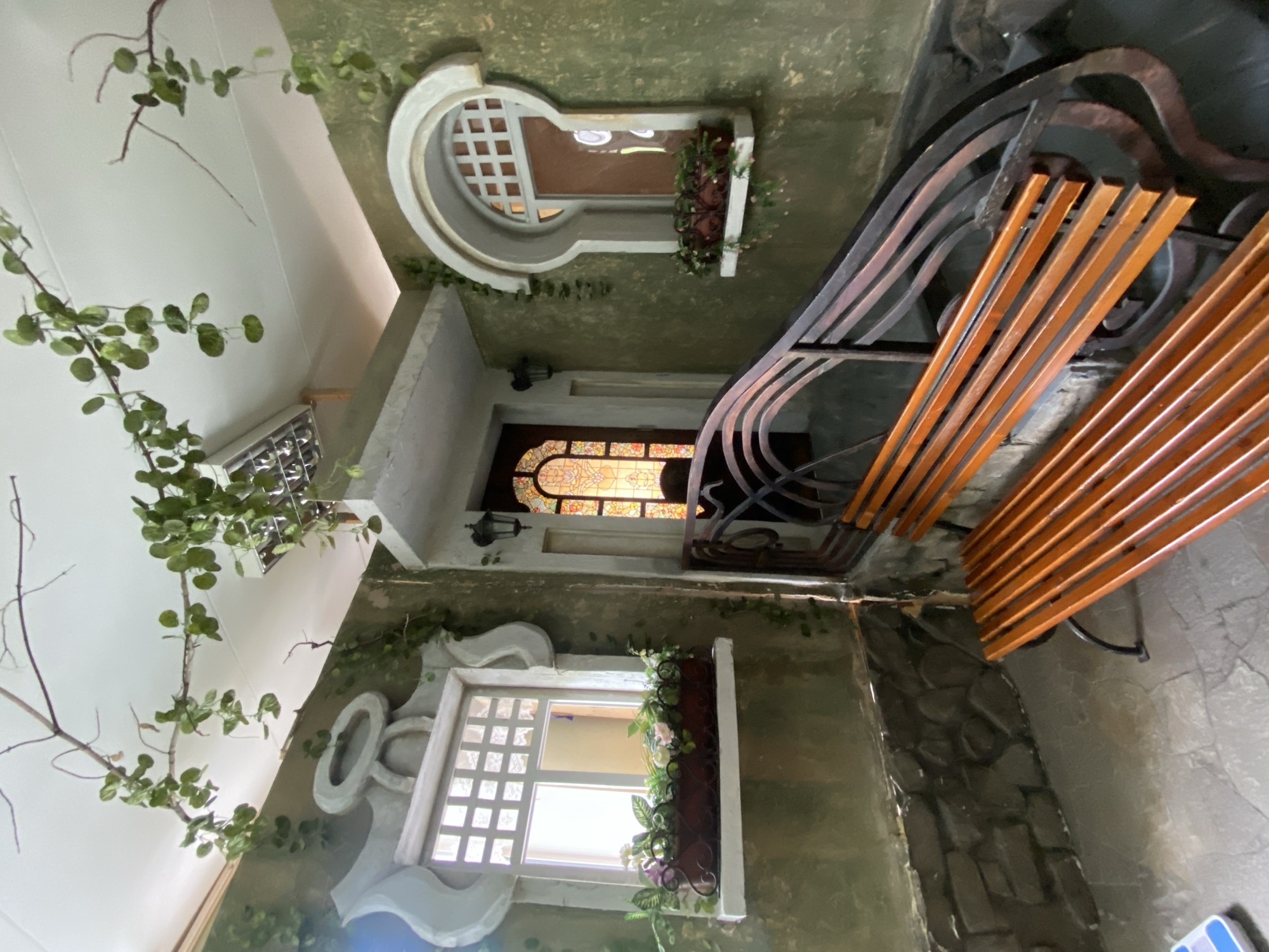 The image shows an interior in the city of Yekaterinburg, Russia. Two windows are present in the frame, one rectangular with a white frame and one circular with a white frame. Both windows contain planter boxes with flowers and greenery. Ivy adorns the walls. A door with stained glass is visible. A metal railing with wooden slats is in the foreground. The walls have a greenish-brown texture.