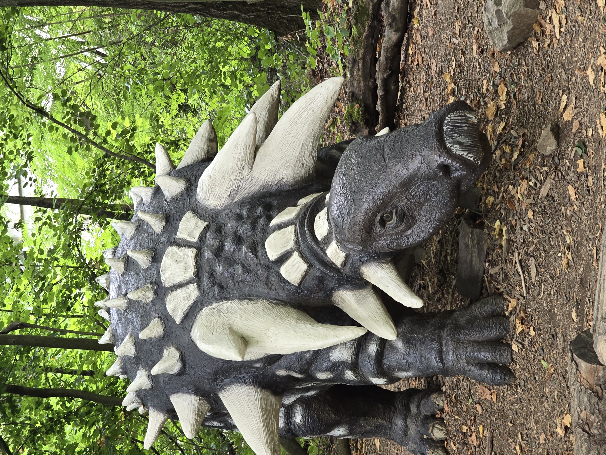 The image depicts a life-sized model of an Ankylosaurus dinosaur positioned in a woodland setting. The model is dark gray with prominent white osteoderms and spikes covering its back and head. Its head is low to the ground, facing the viewer. The surrounding environment consists of trees with dense green foliage and a forest floor covered with dirt, fallen leaves, and some scattered rocks and branches. The lighting suggests an overcast day. The model is situated in a park-like area, possibly part of an outdoor exhibit. This scene is located in the city of Vyškov, Czechia, specifically within the DinoPark Vyškov. There are no people visible in the image, and the dinosaur model is stationary.
