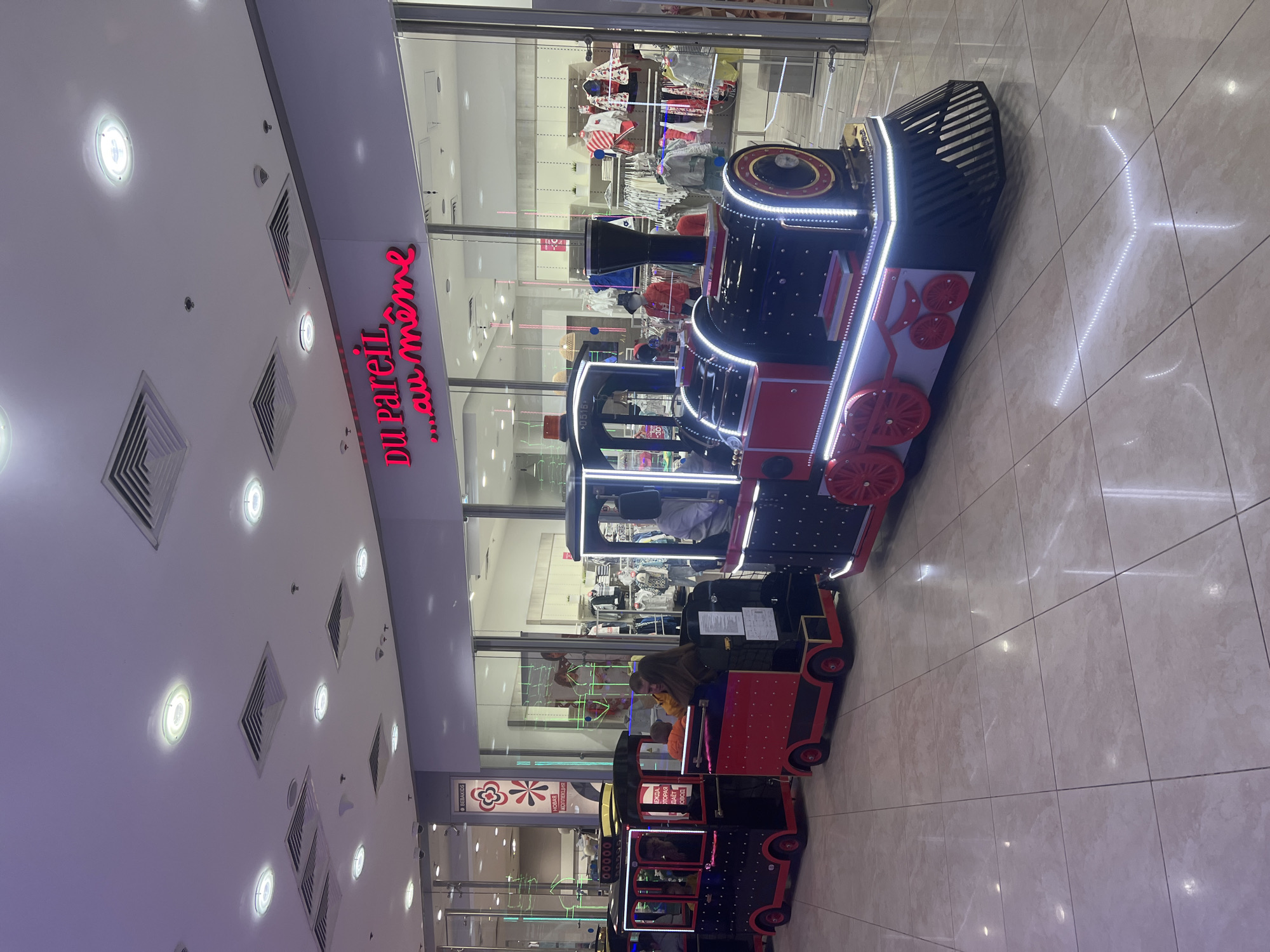 The image captures the interior of what appears to be a shopping mall in Moscow, Russia, as suggested by the Cyrillic text on the storefront signs. A small, brightly colored, motorized train ride, designed for children, is the central focus. Each car is black and red with visible artificial lighting accents. The train is located in front of storefronts, one of which is “Du Pareil au meme”, and another reads “BEBAKIDS” and “новая коллекция”. The ceiling above has an array of recessed lighting fixtures and air vents. The floor is tiled in a light color. In the train's carriages, there are visible people, presumably children and perhaps a caretaker. The environment is brightly lit and spacious, indicative of a modern shopping center.