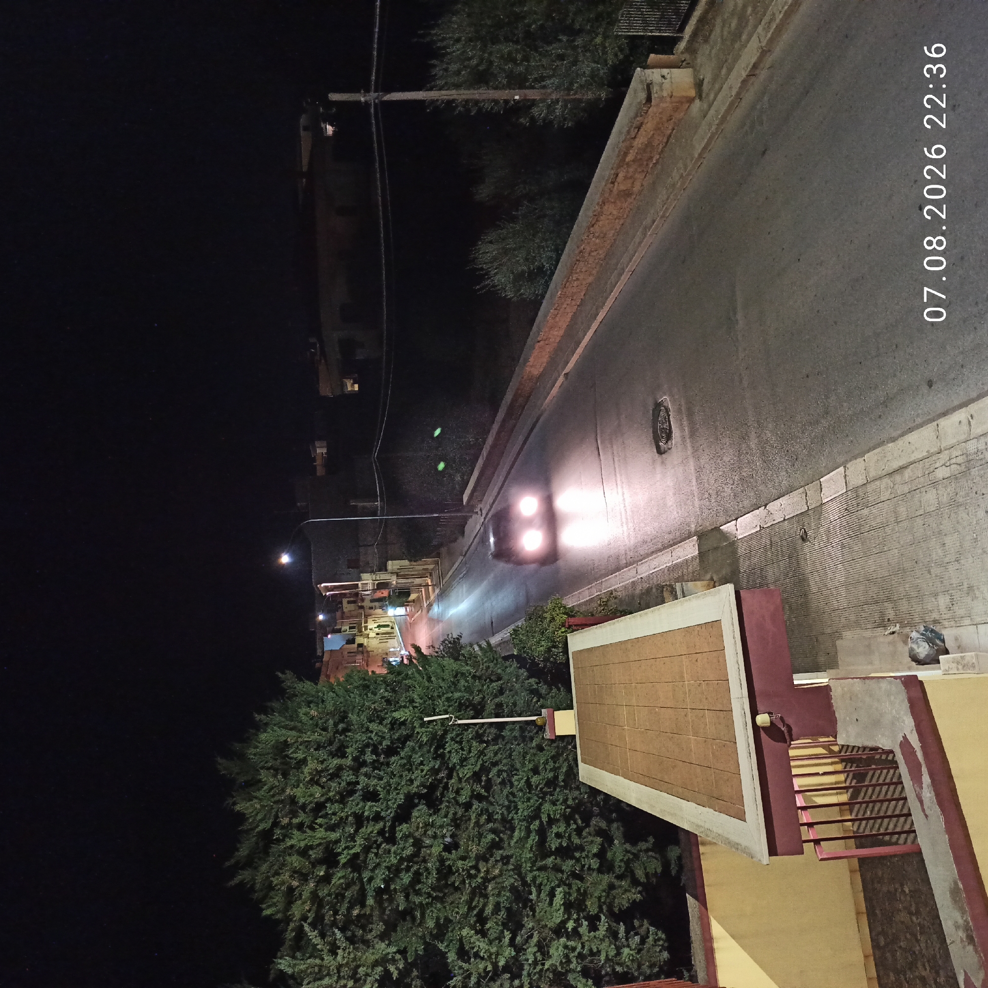 The image captures a street scene at night in Bitonto, Italy, indicated by the timestamp "07.08.2026 22:36". A car with its headlights on is driving down a paved road. The road is flanked by buildings on the left, with trees and vegetation visible. On the right side of the road, there is a stone wall and more greenery. The sky is dark, with some faint stars visible. In the foreground, there is a portion of a building with a yellow wall and a red railing, and a rooftop covered with square tiles. The scene appears to be a residential or mixed-use area within the city.