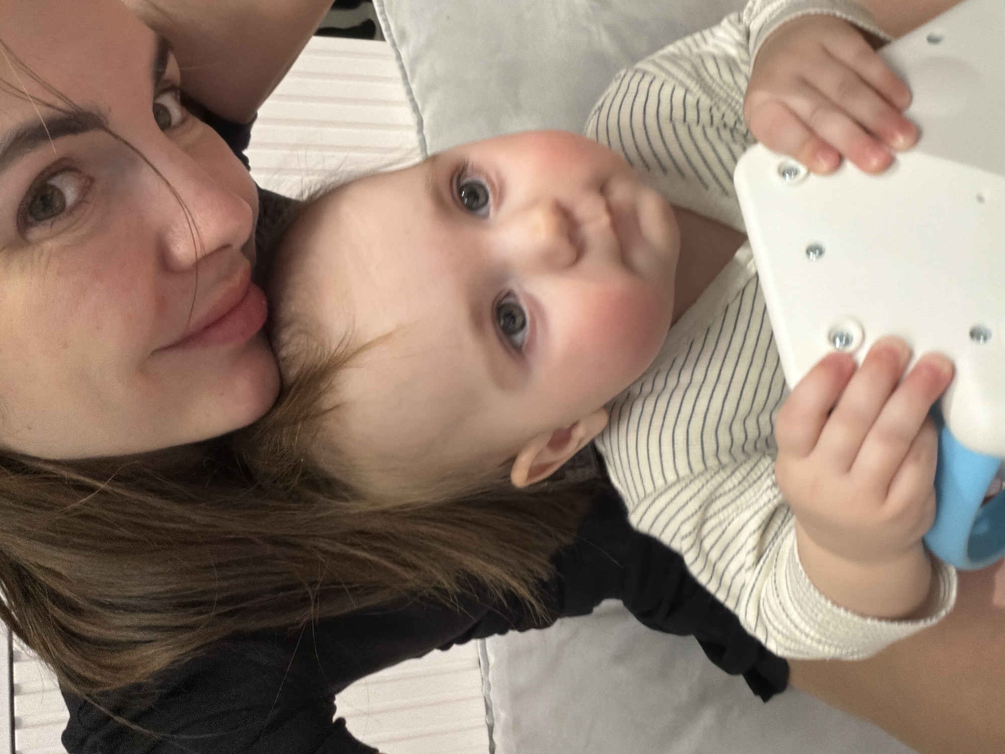 This close-up, selfie-style image captures an adult and a baby sharing a tender moment, likely indoors. The scene suggests a home environment, possibly a bed or soft surface, indicated by the light-colored, subtly textured fabric in the background.
On the left side of the frame, a partial view of an adult woman's face is visible. She has brown hair, dark eyes, and her lips are gently pursed as she looks towards the camera. Her dark clothing is also partially seen.
Occupying the center and right of the frame is a baby, whose face is fully visible and looking directly at the camera. The baby has fair skin with rosy cheeks, light blue or grey eyes, and wispy light brown hair. The baby is dressed in a light-colored outfit with thin dark vertical stripes. The baby's small hands are holding a white, rectangular object, possibly a toy or part of a device, which has several small metallic screws or fasteners on its surface. A light blue, curved component is also visible attached to this object.
The overall impression is one of warmth and affection between the adult and the child. There is no visible text in the image.