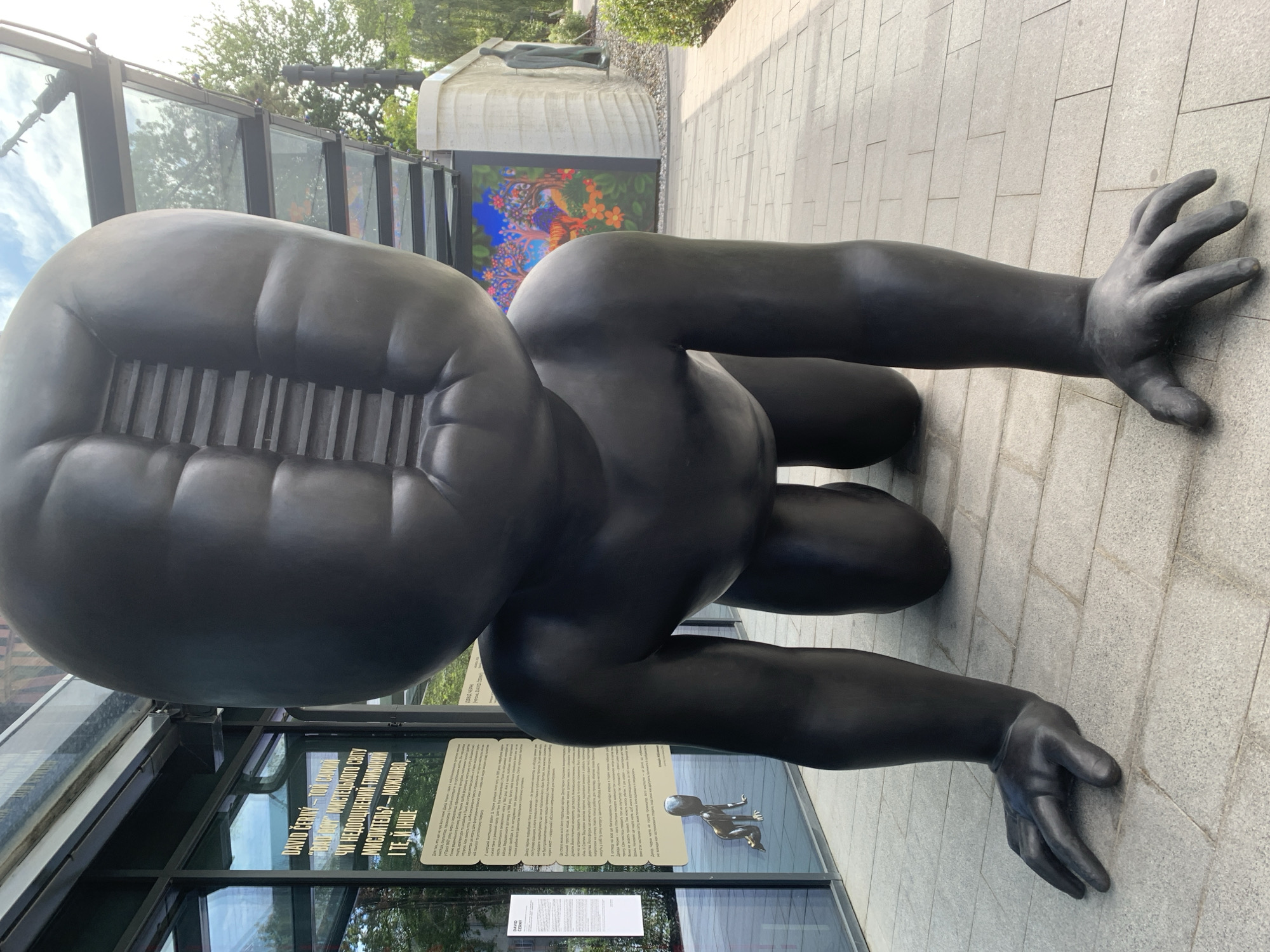 The image shows a large, dark sculpture of a crawling baby with an unusual, grooved opening on its head, positioned outdoors on a paved walkway. Behind the sculpture, there is a glass-fronted building with informational signs displayed on the glass, one of which is partially visible and lists "DAVID CERNY" and "BAD BOY" in English. The sculpture appears to be a public artwork. In the background, there is a brightly colored mural depicting trees, flowers, and an animal, and further beyond, a white wall with another abstract sculpture of a standing figure. The scene suggests an art installation or museum exterior. The overall environment appears to be a modern urban setting with greenery and pedestrian areas. The style of the building and the signage suggest it could be a contemporary art gallery or exhibition space. The sculpture itself is the central focus, with its textured surface and imposing size.