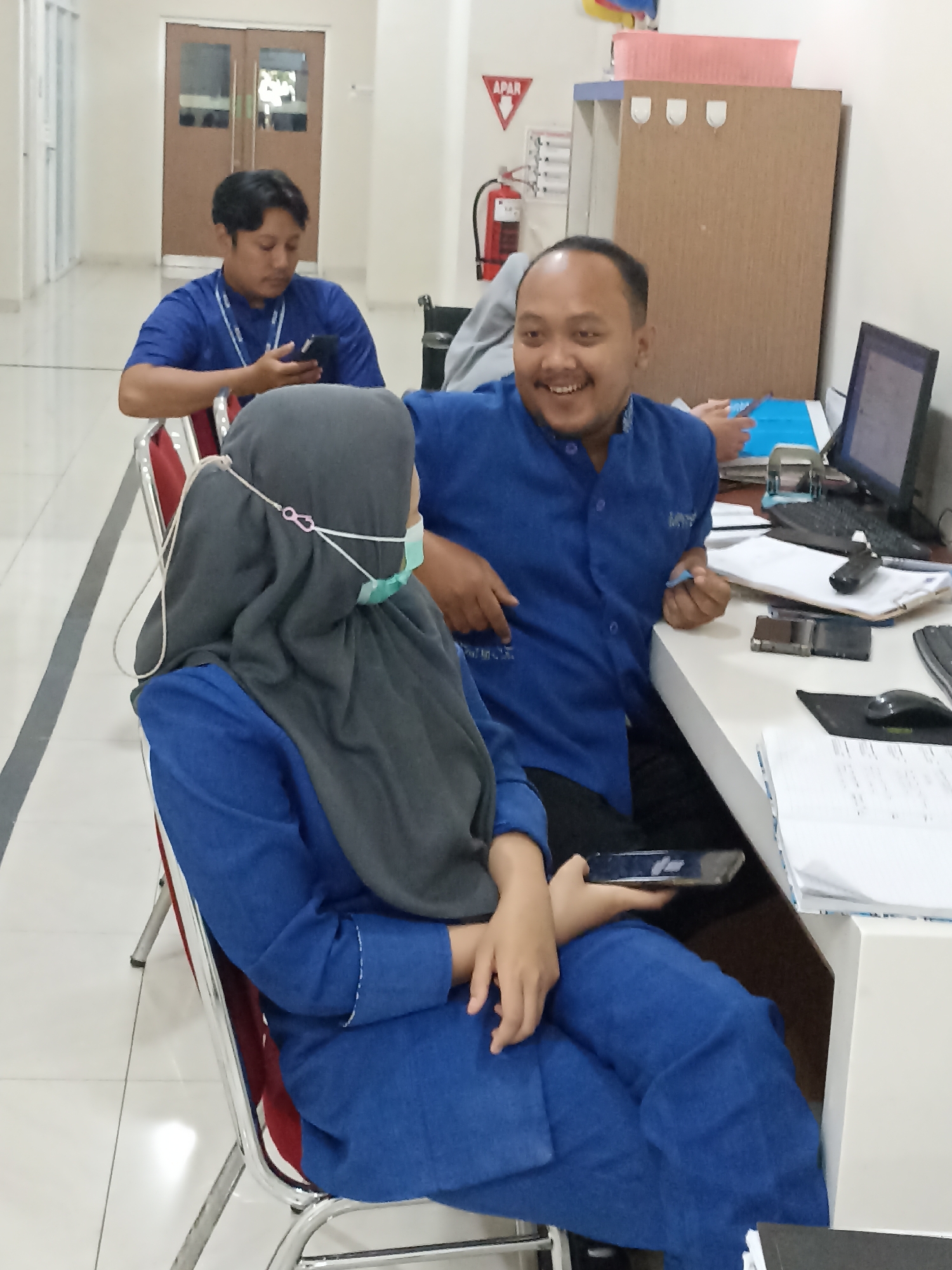 In Beji, Indonesia, three individuals are present in an office setting. Two men and one woman are seated. The man in the foreground, to the right, wears a blue collared shirt and is smiling, looking towards the viewer. He is seated at a desk with a computer monitor, keyboard, and documents. The woman in the foreground, to the left, wears a grey hijab and a light blue face mask, and is looking down at her phone. She is wearing a blue shirt and is seated in a red and silver chair. A third man, in the background, is wearing a blue shirt and is looking at his phone. He is seated in a similar chair to the woman. The environment appears to be a typical office space with filing cabinets, a desk, and chairs. A fire extinguisher is visible on the wall in the background.