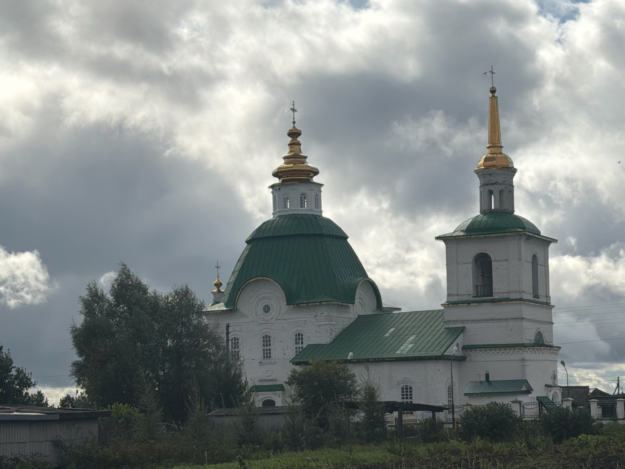 The image shows a white Orthodox church with a green roof and golden domes, located in the village of Preobrazhenka, Russia. The church features multiple architectural elements, including a bell tower and a large dome. It is surrounded by trees and vegetation, with a cloudy sky in the background. The overall scene depicts a religious structure within a rural setting.
