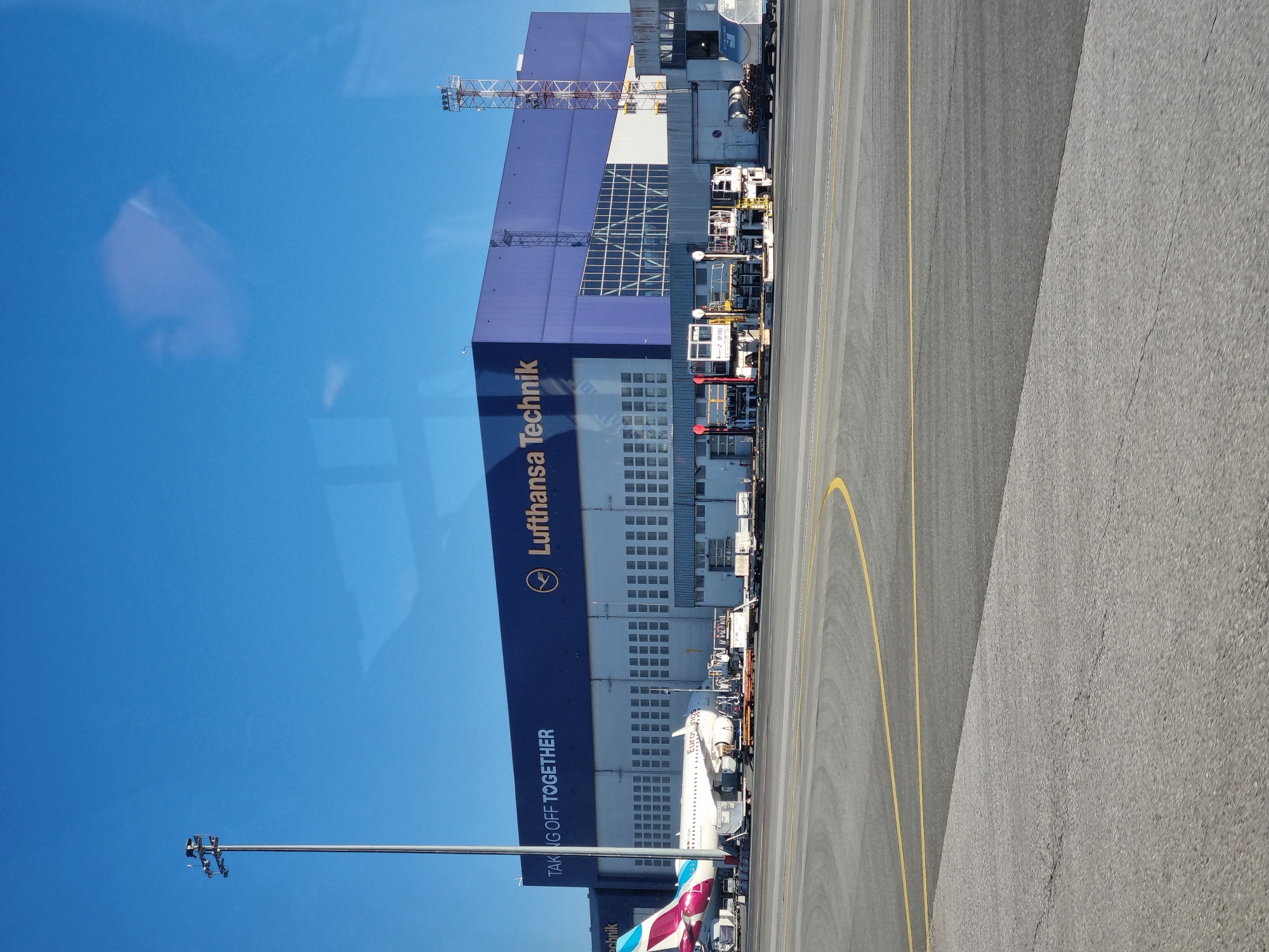 The image shows an exterior view of the Lufthansa Technik building at Sofia Airport, Bulgaria, adjacent to an airport runway. The building is predominantly blue and white, with the Lufthansa Technik logo prominently displayed. Various service vehicles and airport equipment are parked along the side of the building facing the runway. A passenger aircraft with the "Eurowings" logo is parked near the building. The runway is grey, marked with yellow lines. The sky is clear and blue. A light pole is visible in the lower left corner of the image. The image seems to have been taken from inside a vehicle.