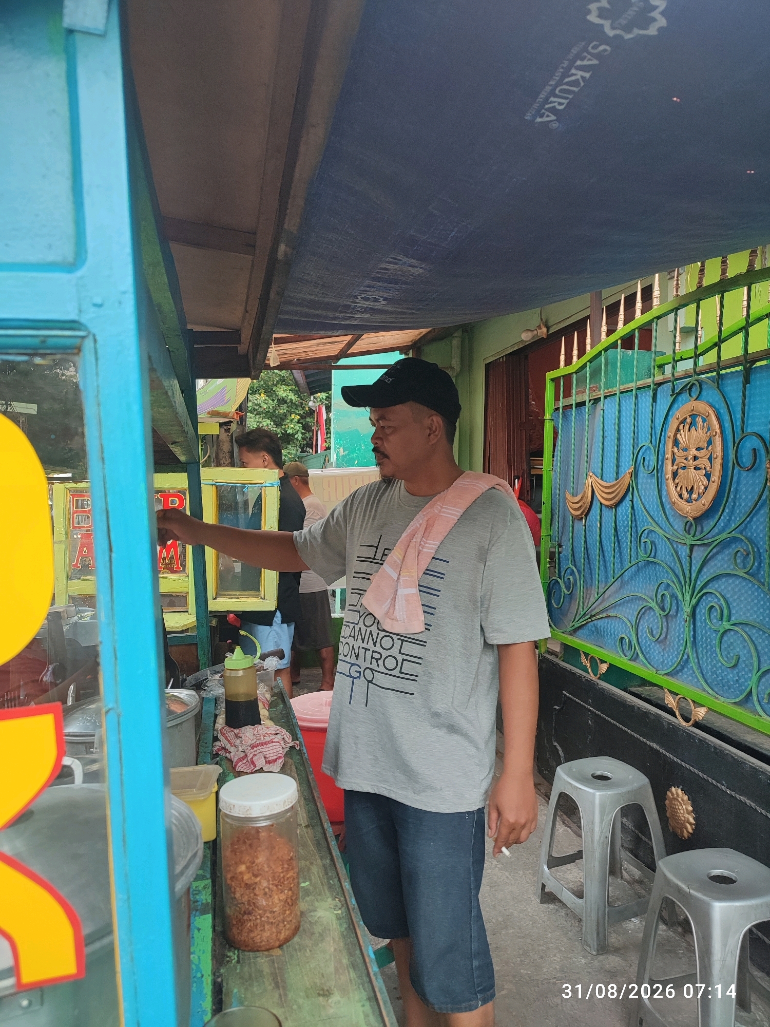 A man in a gray t-shirt and blue shorts stands at a food stall with a blue and green exterior. He wears a black baseball cap and has a pink towel draped over his shoulder. He is holding a cigarette and appears to be serving food. Behind him, there is a green metal gate with ornate gold decorations and two plastic stools. The stall appears to be located in East Jakarta, Indonesia, as indicated by the timestamp on the image. The scene suggests a street food vendor operating in a residential or commercial area.
