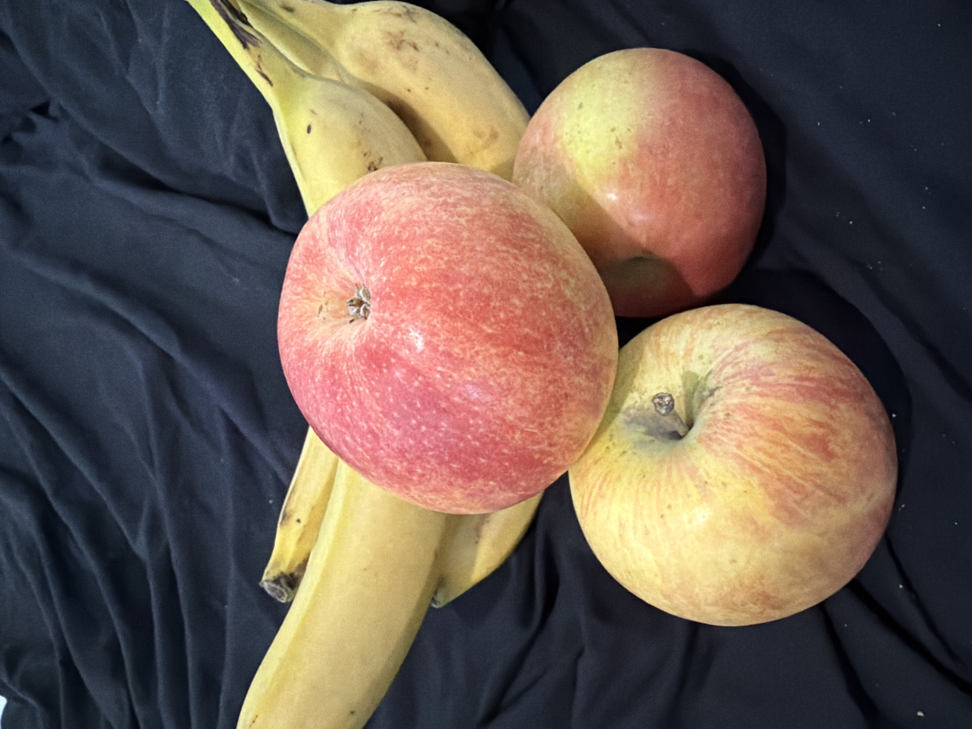 The image displays a still life composition of fruit arranged on a dark fabric. The primary subjects are three apples and a bunch of bananas. The apples are round with a mix of red and yellow coloring, exhibiting a blush of pink in some areas. One apple is prominently in the foreground, centered and facing the viewer. To its right and slightly behind it, another apple is visible, mostly showing its side. A third apple, partially obscured by the others, is positioned to the upper right.
The bananas are a vibrant yellow, with some darker speckles, indicating ripeness. They are clustered together and extend from the upper left corner down towards the center of the image. The dark fabric beneath the fruit is wrinkled, creating shadows and adding depth to the scene. The lighting suggests an indoor setting, possibly a studio or a kitchen counter, with soft light illuminating the fruits. There is no discernible text in the image. The scene is static, with no activity taking place other than the arrangement of the fruit for the photograph. The overall mood is simple and natural, focusing on the textures and colors of the produce.