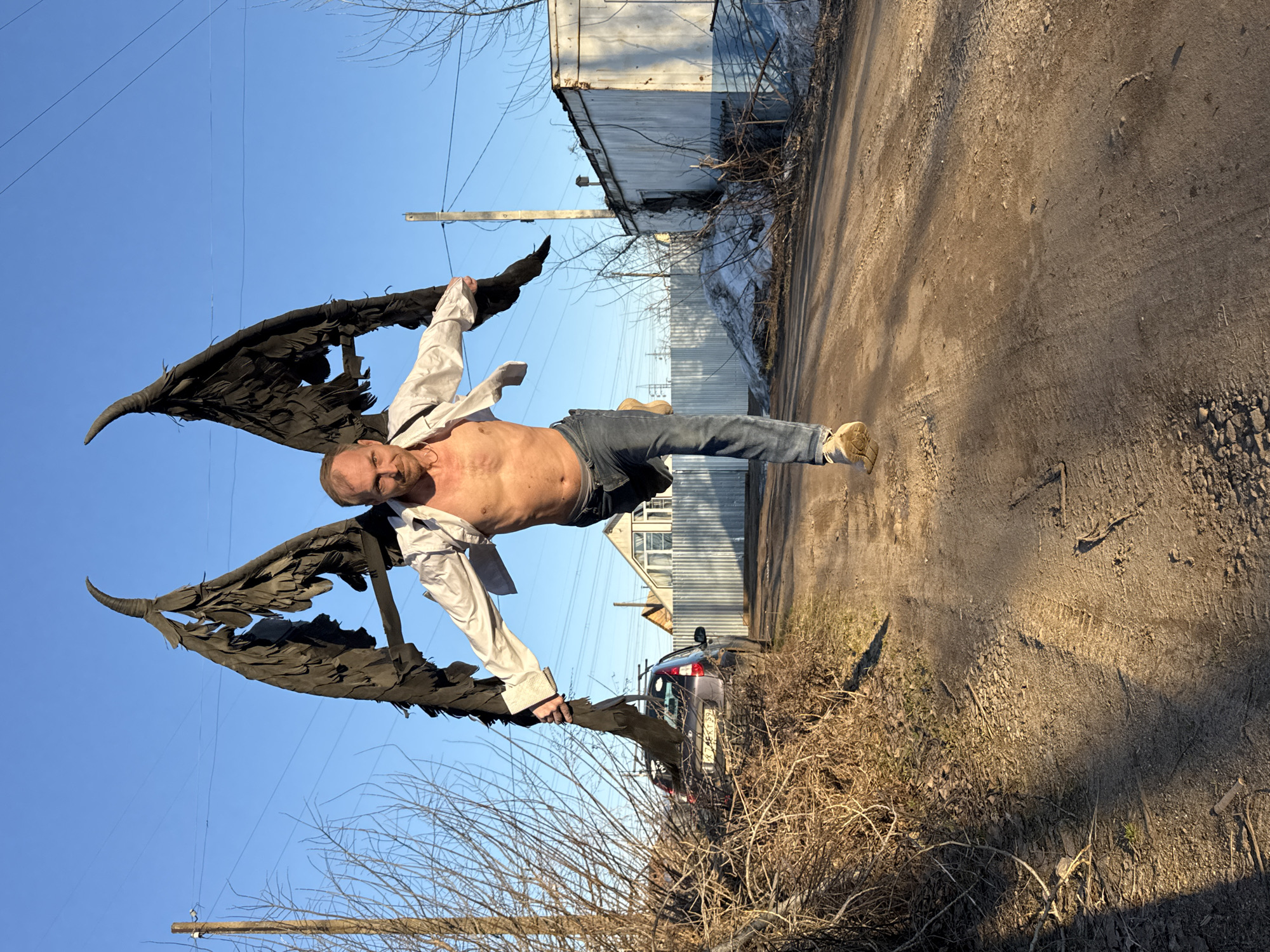 A man stands in a vacant lot in Chaikovsky, Russia, wearing jeans, tan shoes, an open white shirt and large black wings. The background includes a dirt road with tire tracks, metal buildings, a car, bare trees, and a clear blue sky. The man is bald, shirtless, and wearing glasses. He is holding the wings out to each side, apparently striking a flying pose.