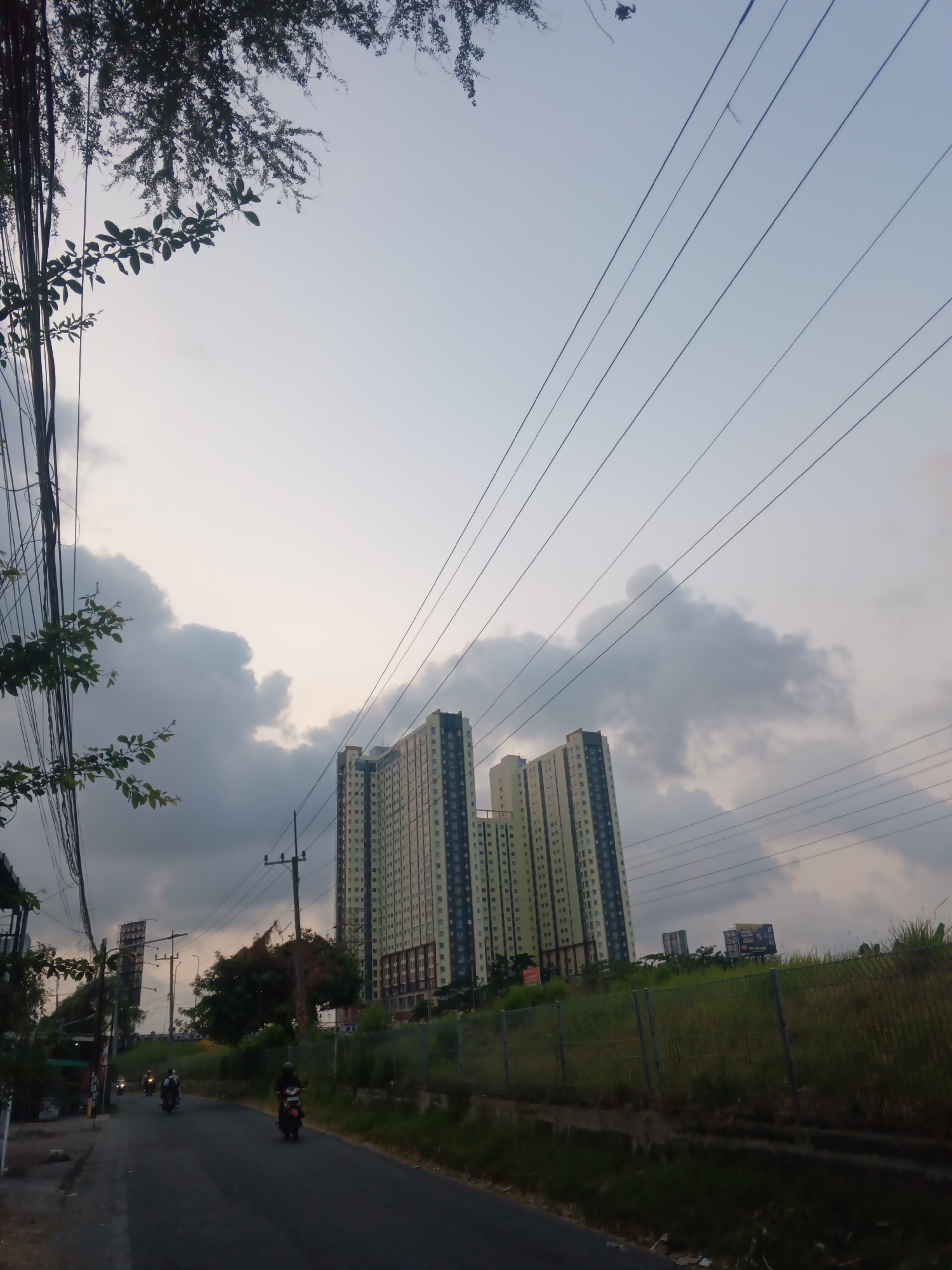 This is a photograph taken on a street in Surabaya, Indonesia, likely in the afternoon or early evening, given the lighting conditions. The scene depicts a road that curves slightly to the left, with a few motorcycles being ridden by individuals wearing helmets. On the left side of the road, there are buildings and trees, with numerous electrical wires overhead. To the right of the road, there is a grassy embankment with a fence, and in the background, two tall apartment buildings are visible. The sky is overcast with large, dark clouds.
