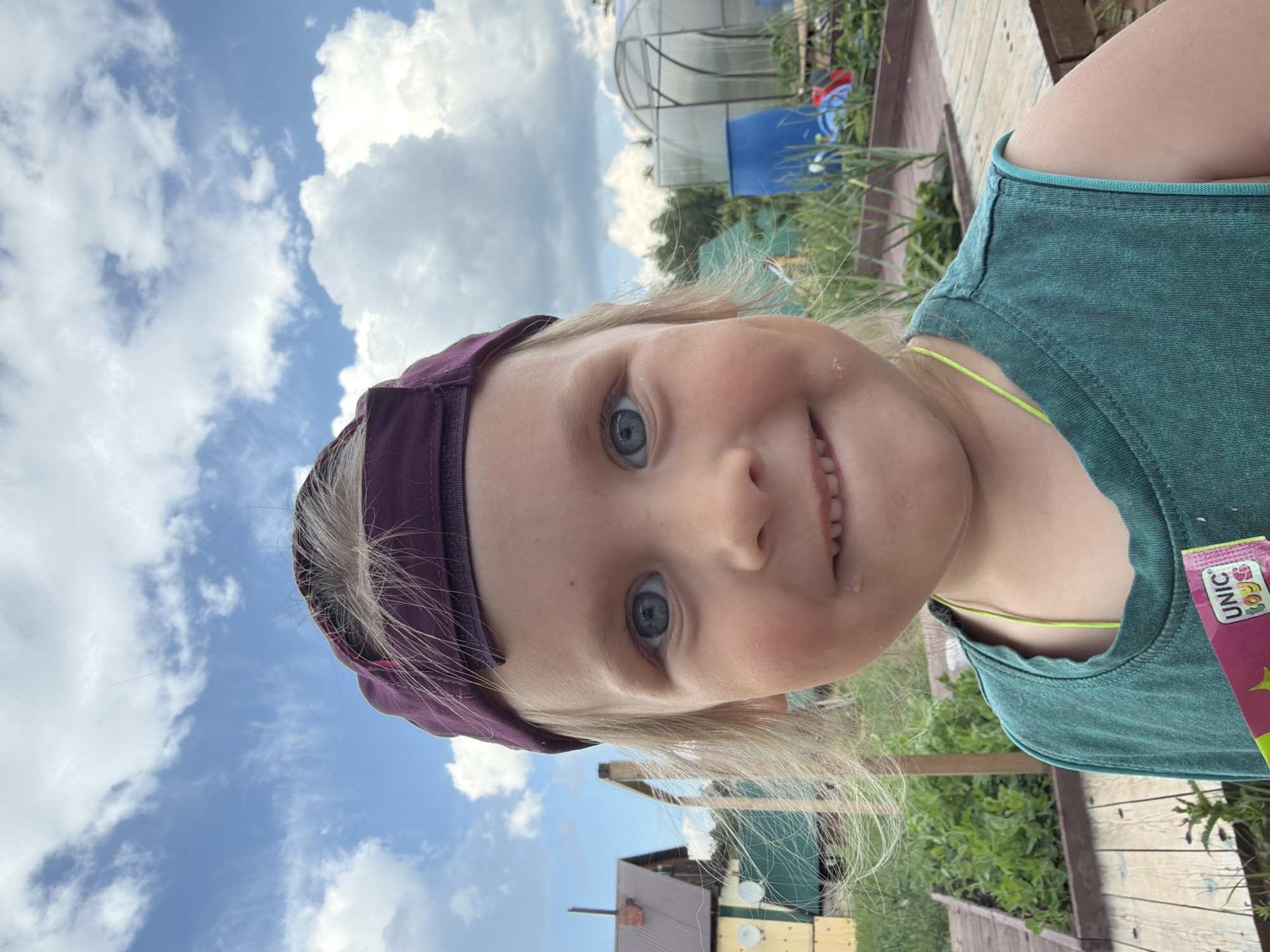 A close-up outdoor portrait shows a young child wearing a purple baseball cap and a teal t-shirt, standing in what appears to be a dacha or garden setting. The child's head is tilted slightly, and they are looking directly at the camera with a wide smile. Their blonde hair is visible around the sides of the cap, and their blue eyes are prominent. A small, pink and yellow package, possibly a candy wrapper, is held in the lower right corner of the frame, partially obscuring the child's t-shirt.
In the background, several structures and elements suggest a rural or suburban environment, consistent with a dacha in Chaikovsky, Russia. To the left, a small wooden building with a dark roof and yellow siding is visible. A greenhouse structure with a clear, curved roof is situated in the background to the right. Various green plants and garden beds are also present, along with wooden walkways and a large blue barrel. The sky is a bright blue with scattered white clouds. The overall lighting indicates a daytime setting, likely during fair weather.