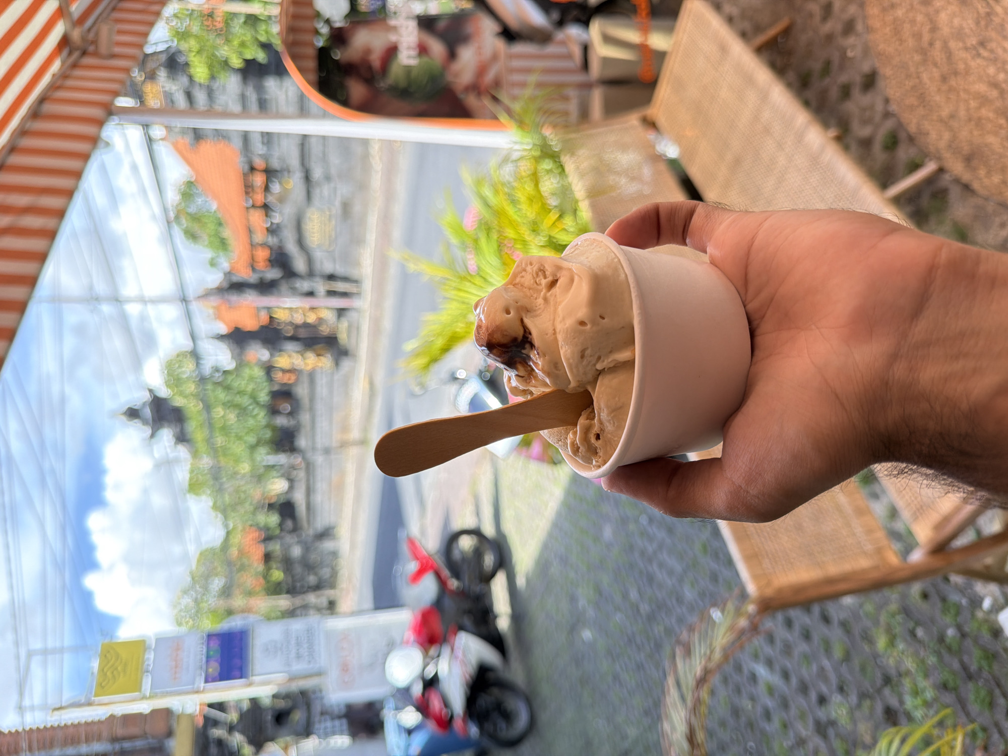 A first-person perspective shot shows a fair-skinned hand holding a small white paper cup filled with light brown ice cream. A small wooden spoon is inserted into the ice cream. Behind the cup and hand, a blurred street scene is visible, featuring a parked red and white scooter. In the background, there are signs, buildings with traditional Balinese architecture, and trees under a blue sky with clouds. Wicker furniture is also partially visible in the foreground. The location appears to be North Kuta, Indonesia, based on the architectural style and general scenery.