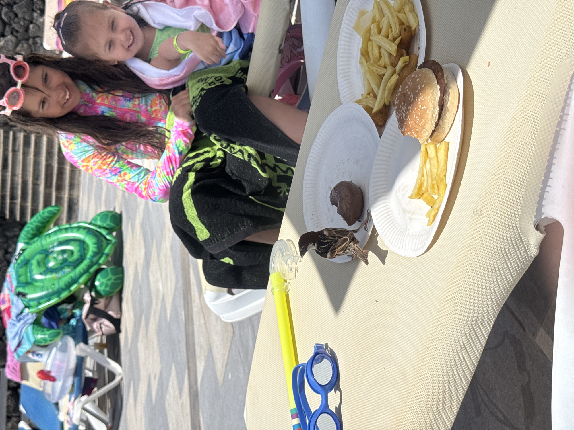 On a sunny day in Puerto Del Carmen, Spain, two young girls are sitting on a beach chair or sun lounger, with plates of food and beach accessories nearby. One girl wears a pink and multi-colored long-sleeved shirt and has pink sunglasses on her head; the other girl has a green shirt and a white towel draped over her shoulders. A plate of french fries and a hamburger are visible. A small brown bird is perched near a plate with a brown meatball on it. A yellow and blue snorkel set lies on the lounger. A green inflatable turtle toy sits on the ground next to the lounger. The environment includes paved steps leading up to a darker surface.