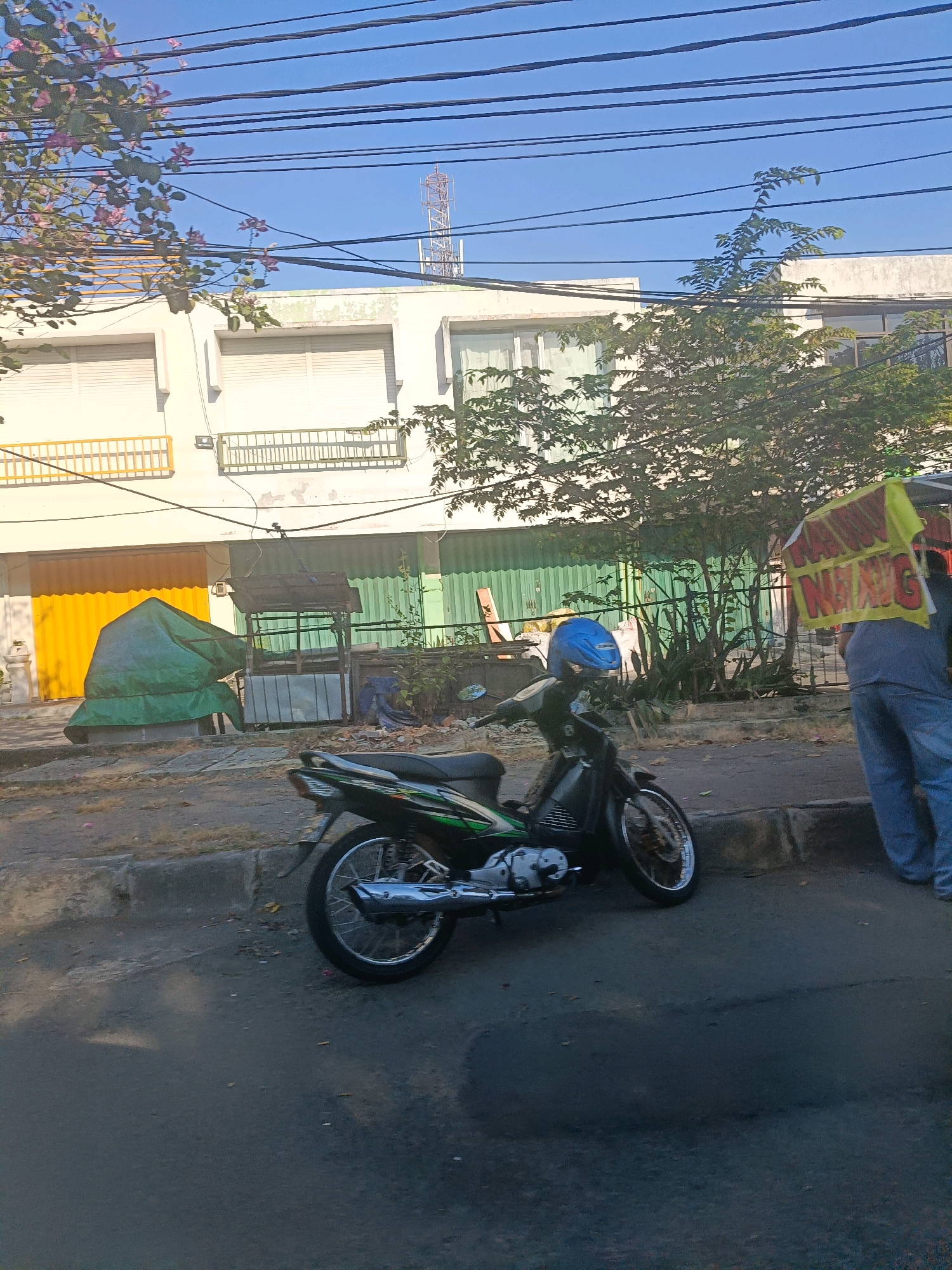 The image depicts a street scene in Surabaya, Indonesia, with a focus on a parked motorcycle and a person holding a sign. The motorcycle, a black and green model with a blue helmet placed on its seat, is positioned on the side of the road, next to a concrete curb. The background features a two-story building with closed shutters and several trees. To the right of the motorcycle, a person wearing blue jeans and a gray shirt is partially visible, holding a yellow sign with red lettering that reads "NAWANG SARI." The street is paved with asphalt, and there are some debris on the ground. Overhead, multiple power lines stretch across the frame. The overall impression is a snapshot of everyday urban life in Surabaya.