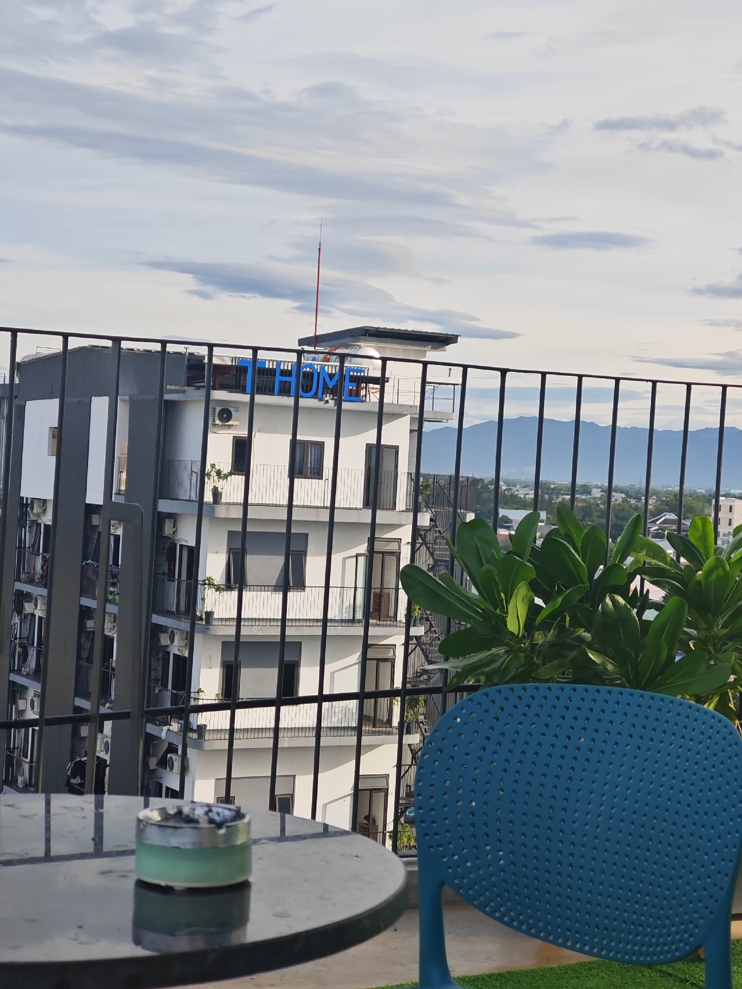 The image captures a view from a balcony overlooking a building with a prominent "HOME" sign in blue letters on its rooftop. The building, located in Đà Nẵng, Vietnam, features multiple floors with balconies and air conditioning units. In the foreground, a round table holds an ashtray with cigarette butts. A blue chair is partially visible, facing away from the camera. Lush green plants are positioned behind the railing, and in the distance, mountains form a hazy backdrop under a cloudy sky. The scene appears to be an outdoor seating area, possibly a cafe or residential balcony.