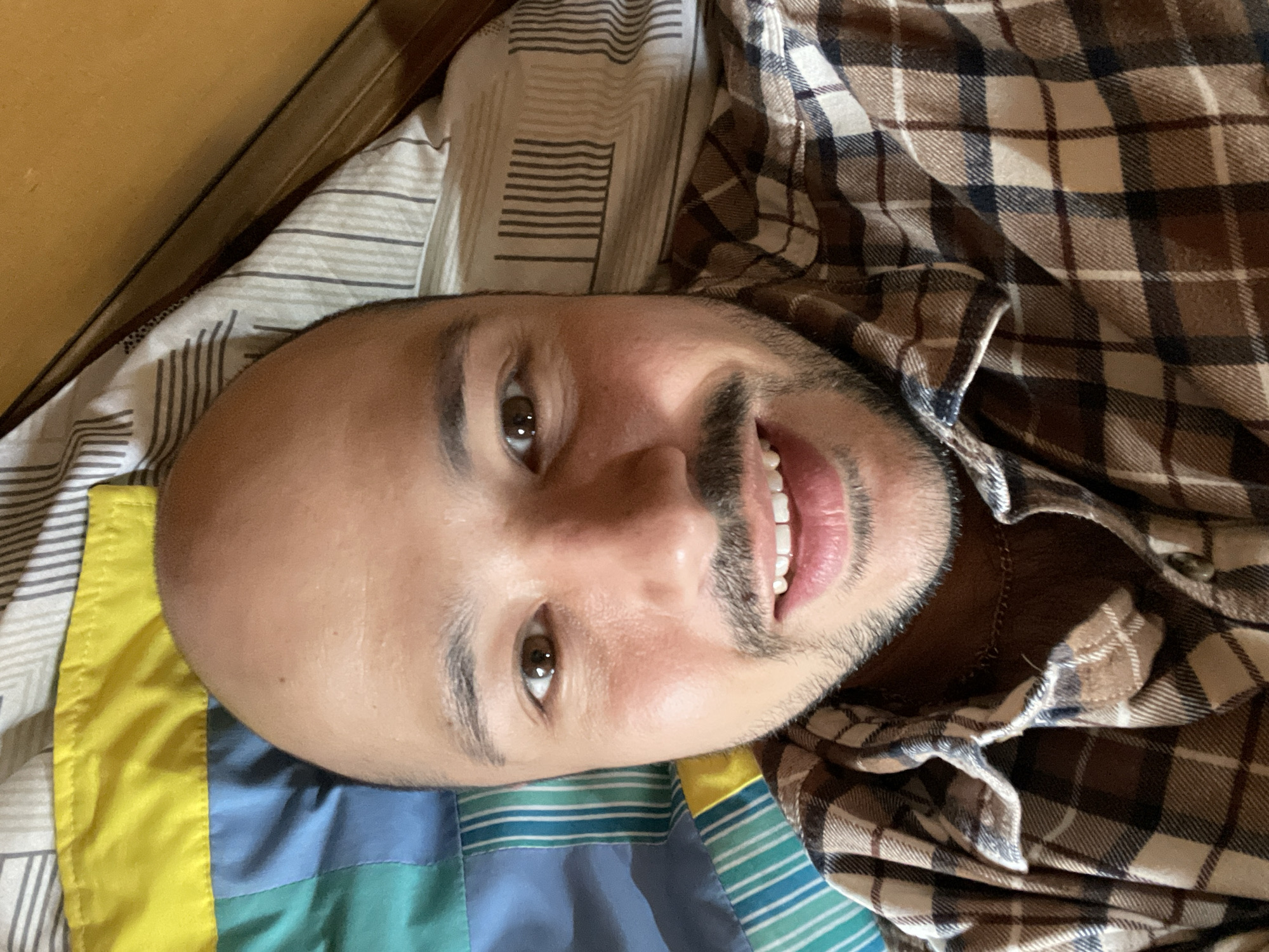 A selfie of a bald-headed man with brown eyes and a mustache lying on a bed. He is wearing a brown and tan plaid shirt and smiling. The bed has a patterned pillow and a colorful blanket. The location of the image cannot be determined from the visual data.