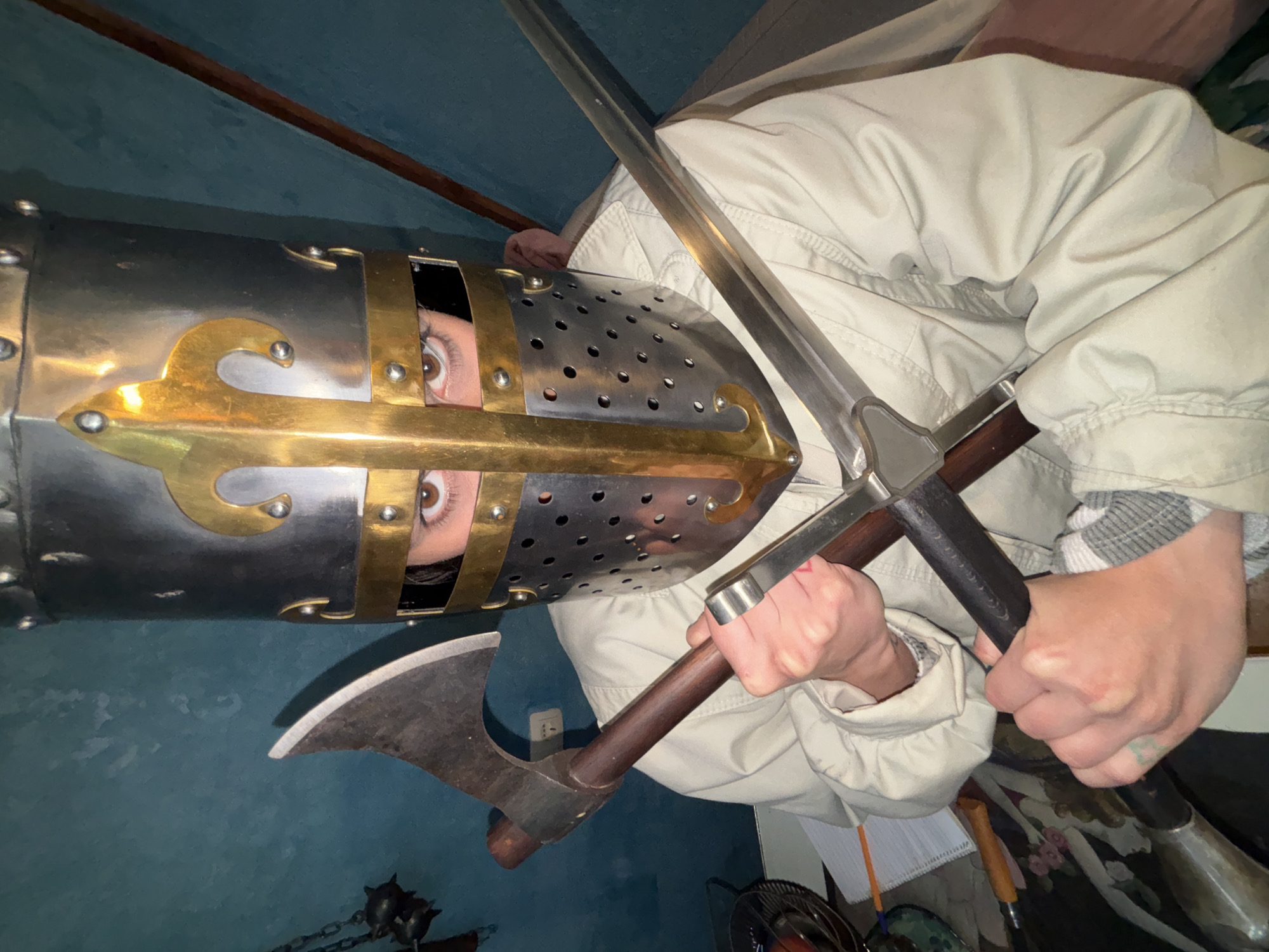 A person is wearing a medieval crusader helmet and holding both a sword and an axe. The helmet is silver with gold accents and has eye slits. The sword is silver, and the axe has a brown wooden handle. The person is wearing a light-colored jacket. The background is dark blue. The scene appears to be indoors. The location is not discernible from the image.