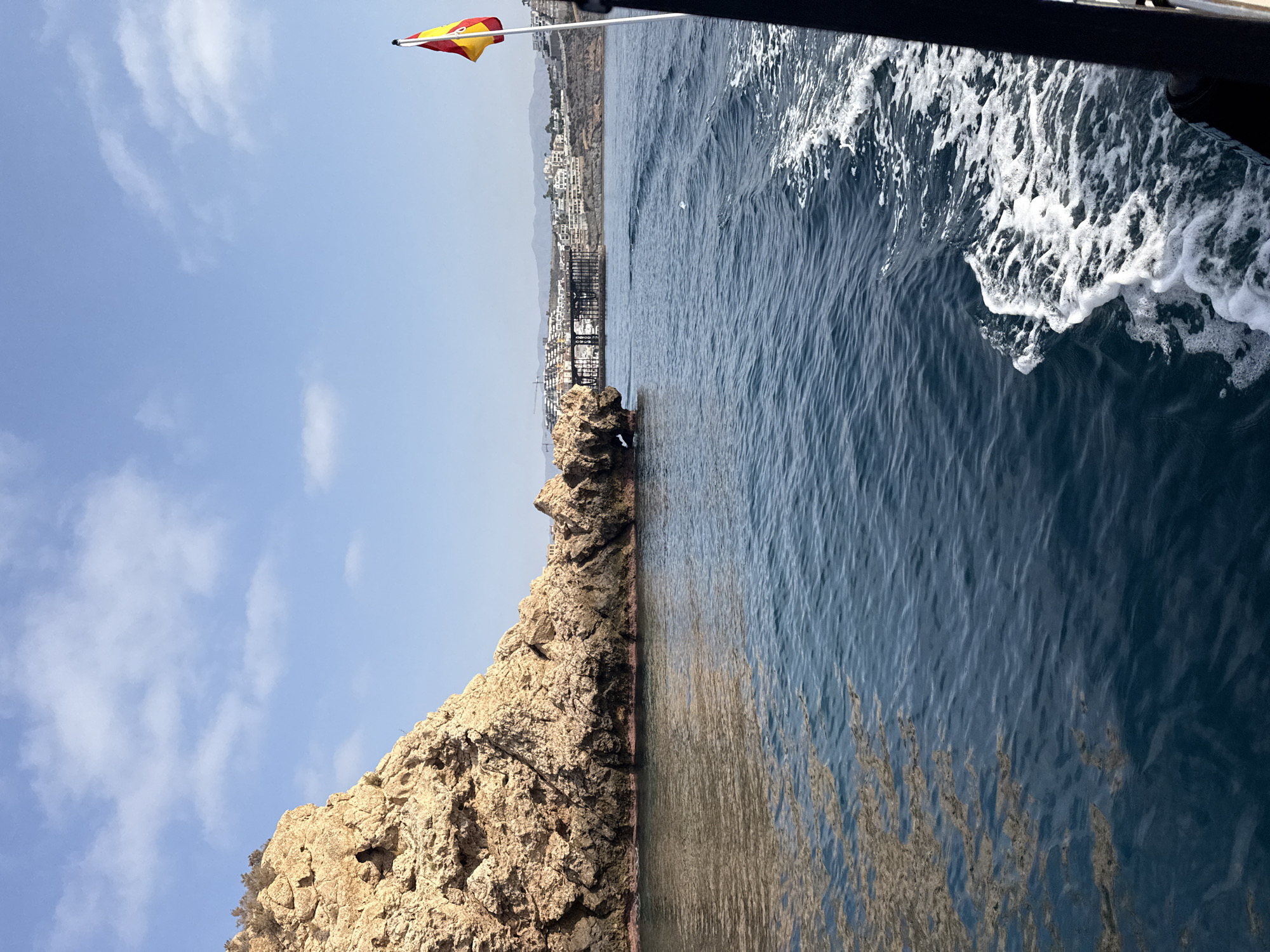 This is a medium-angle, exterior, daytime, eye-level shot of a rocky outcropping in the sea, viewed from a boat moving away from it. The water is deep blue and choppy, with white foam trails indicating the boat's wake on the right. A Spanish flag flutters from a pole on the right side of the frame. In the background, a town with white buildings is visible along a coastal hillside, with mountains beyond. A dark, industrial-looking structure, possibly a pier or bridge under construction, is situated between the rocky outcropping and the town. The sky is a light blue with scattered white clouds. The rocky outcropping is a prominent feature, with rough, textured surfaces and dark shadows at its base.
