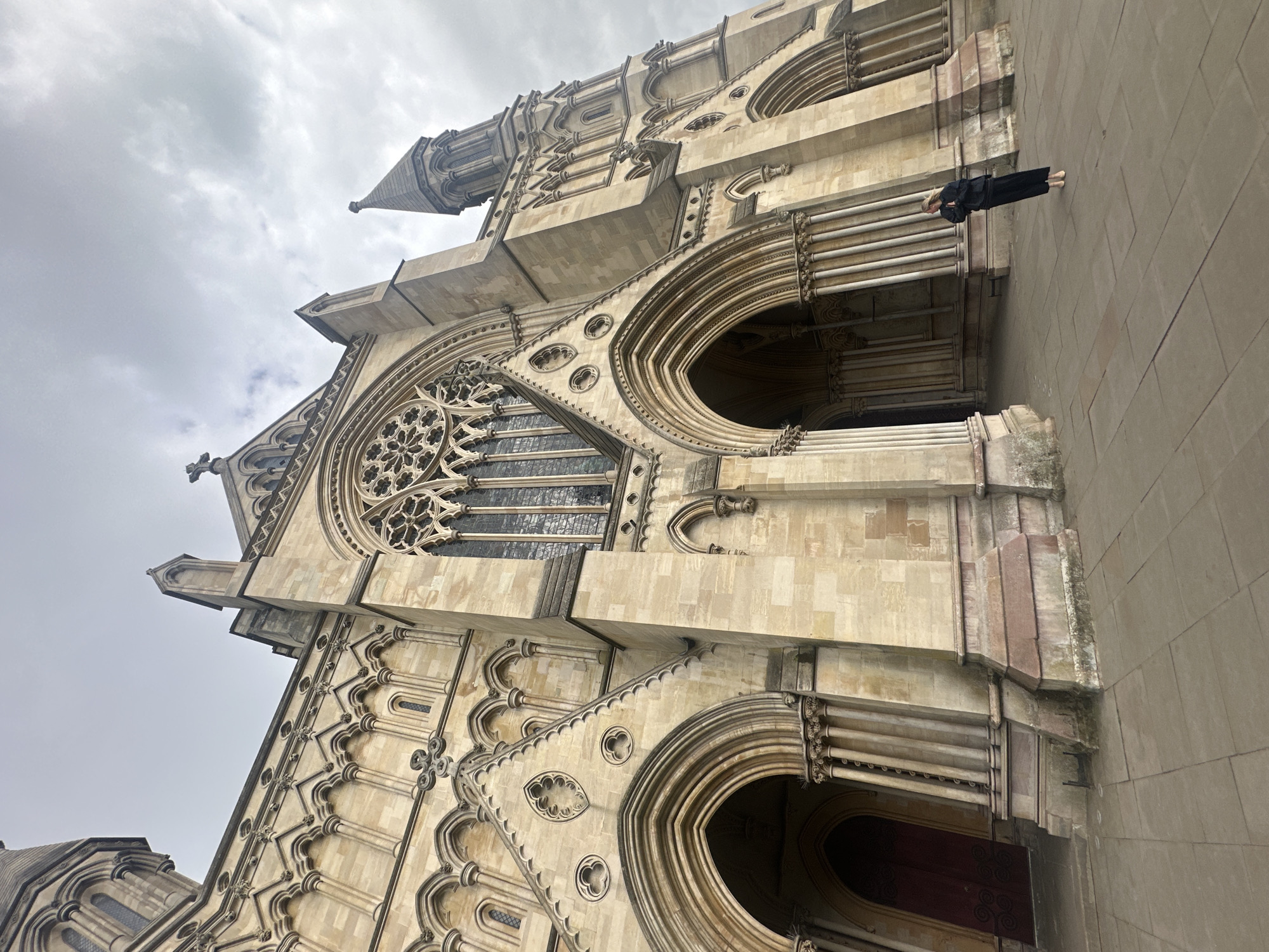 The photograph depicts the exterior of St Albans Cathedral in St Albans, United Kingdom. The cathedral is constructed of light-colored stone with intricate architectural details, including arched windows, rose windows, and towers. The sky is overcast with gray clouds. A person dressed in dark clothing is positioned on the right side of the frame, appearing to lean against the wall of the cathedral.