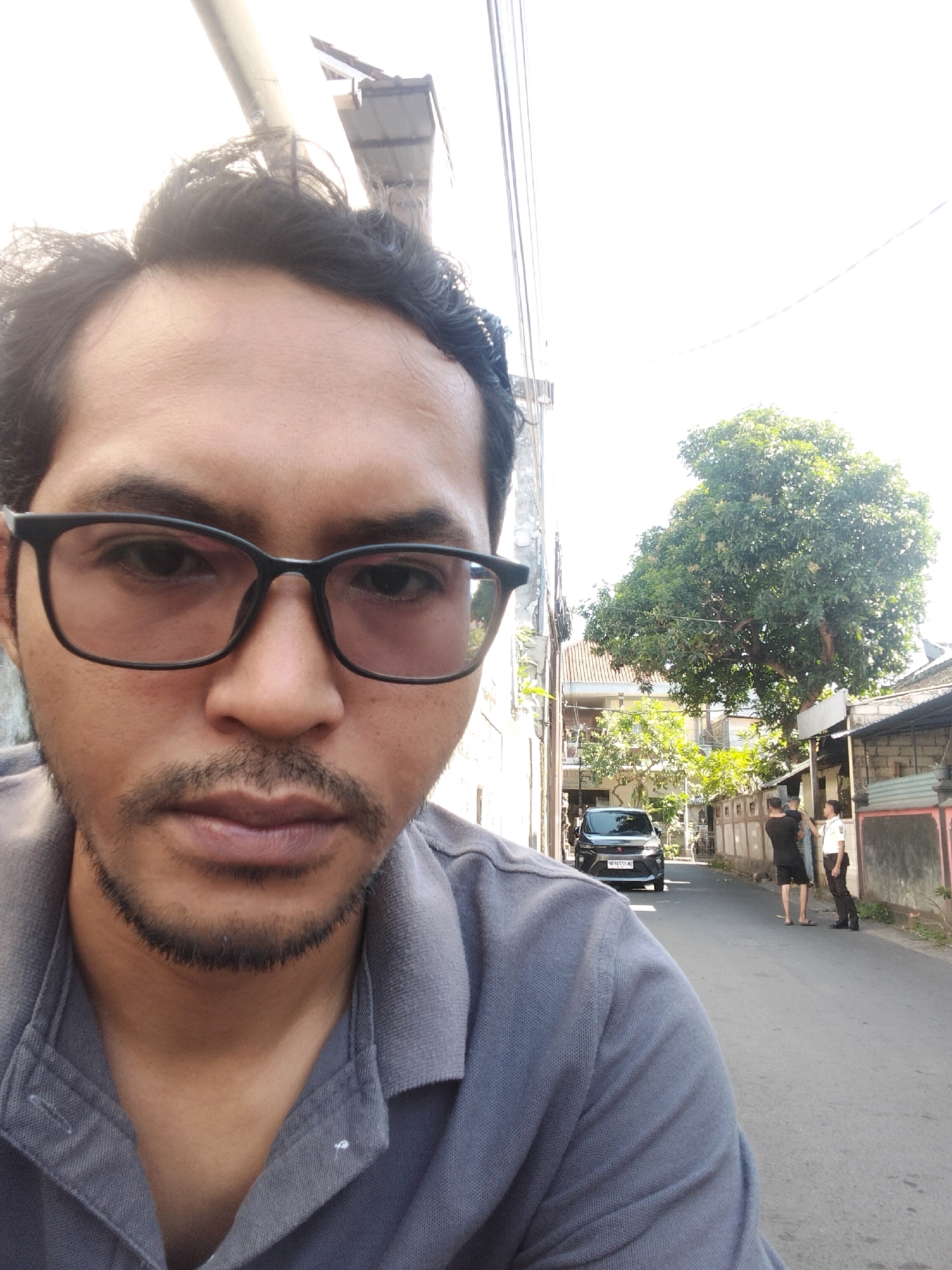 A selfie of a man in his 30s wearing glasses and a grey polo shirt, with a mustache and goatee, is taken outdoors in an alleyway in Kuta, Indonesia. The man's face occupies the foreground and is in sharp focus. In the background, a black car is parked on the left side of the street, and two people are standing on the right side of the street, one holding a child. A large tree with green foliage is visible in the background, along with houses and a building with a tiled roof. The sky is bright and washed out, indicating a sunny day. The man is looking directly at the camera.