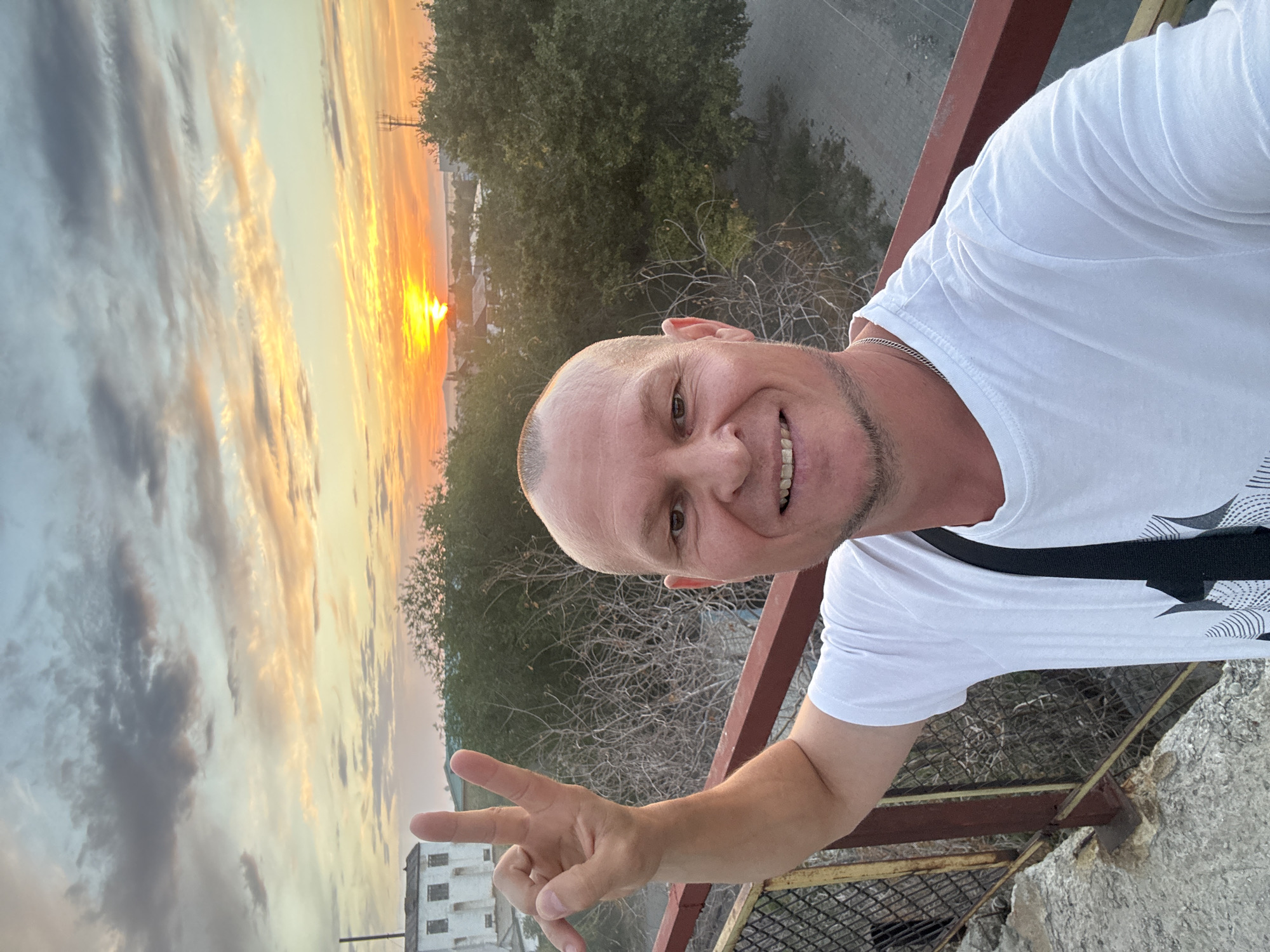 A man with a shaved head and a beard smiles and makes a peace sign at the camera, holding his right arm up. He is wearing a white t-shirt with a black strap from a bag across his chest. Behind him, a sunset illuminates the sky with orange and yellow hues, casting a glow over a city in the distance. Trees and a white building are visible in the midground, and a fence with red and yellow bars runs along the foreground. The scene appears to be an outdoor setting, possibly on a rooftop or elevated vantage point, overlooking the city of Charsk, Kazakhstan.