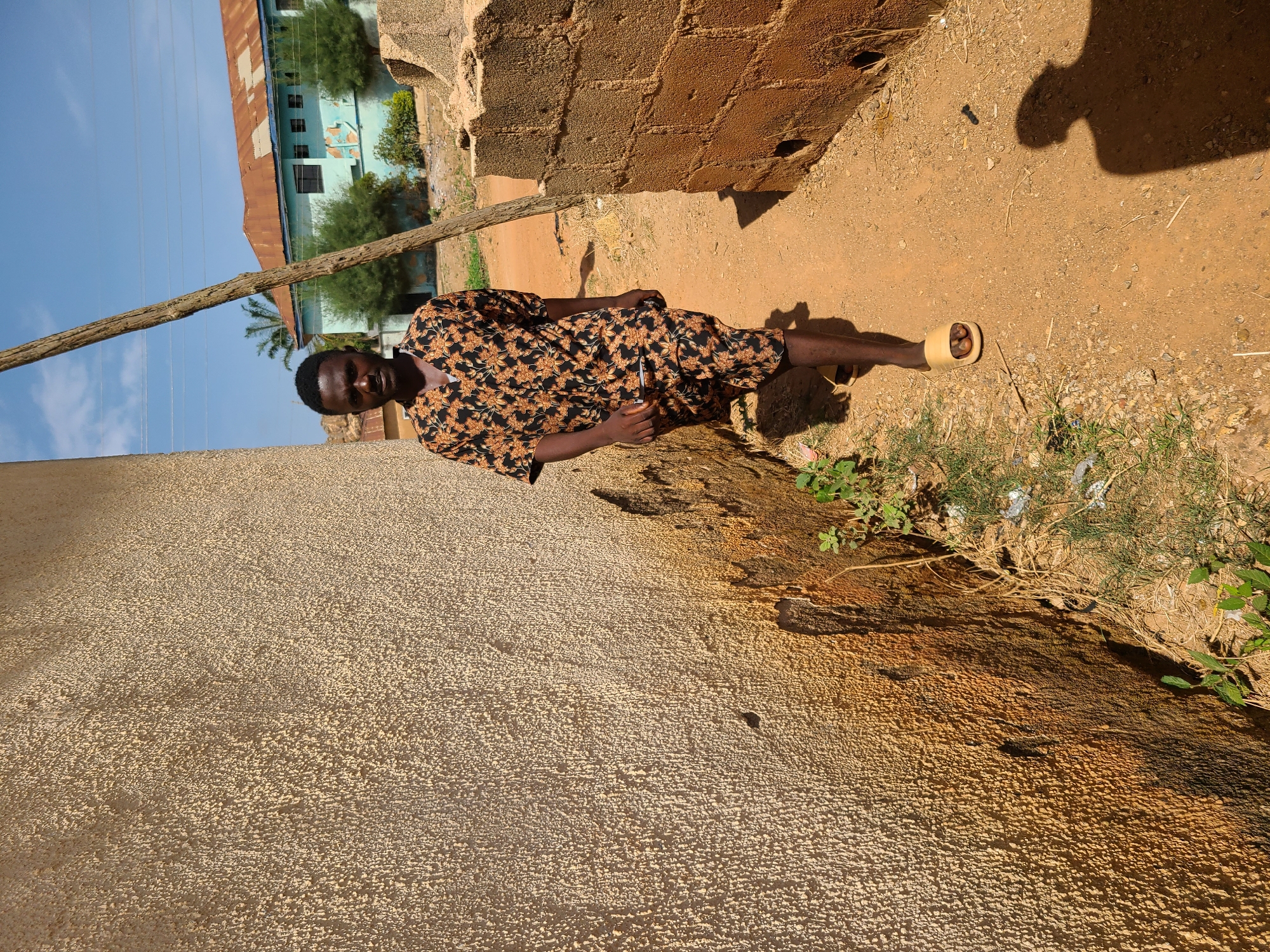 A man in a floral shirt and shorts is lying on the ground in Jos, Nigeria. He is wearing a sandal on one foot. He is next to a weathered wall and a dirt path. There is a wooden pole and a building in the background under a clear blue sky. The ground is littered with small pieces of trash and vegetation. The shadow of a person is cast onto the dirt path.