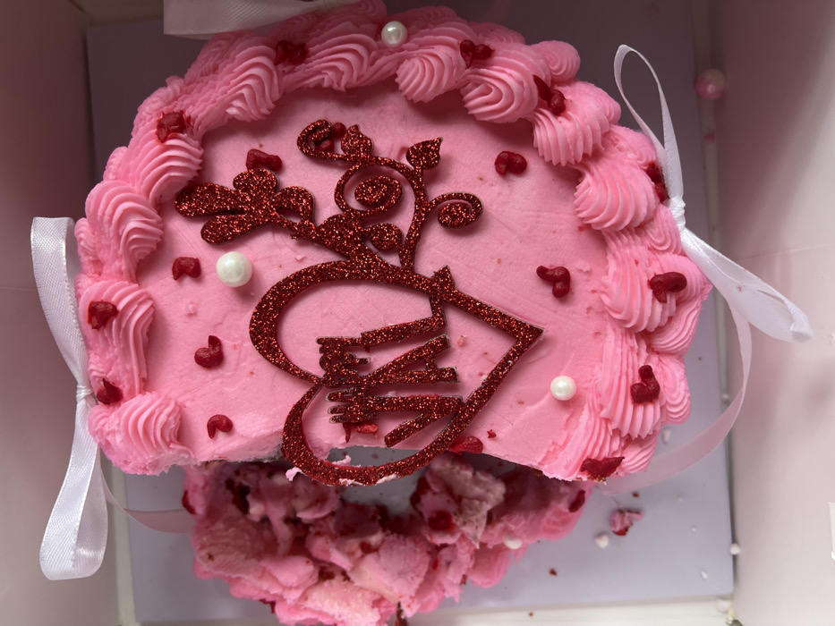 A pink cake with pink frosting sits in a white box. The cake is decorated with red glittery designs. Some of the cake has been broken off, revealing its soft texture. White ribbons adorn the sides of the cake. The cake's surface is adorned with small red hearts and white pearl-like decorations. The frosting is swirled elegantly around the edges.