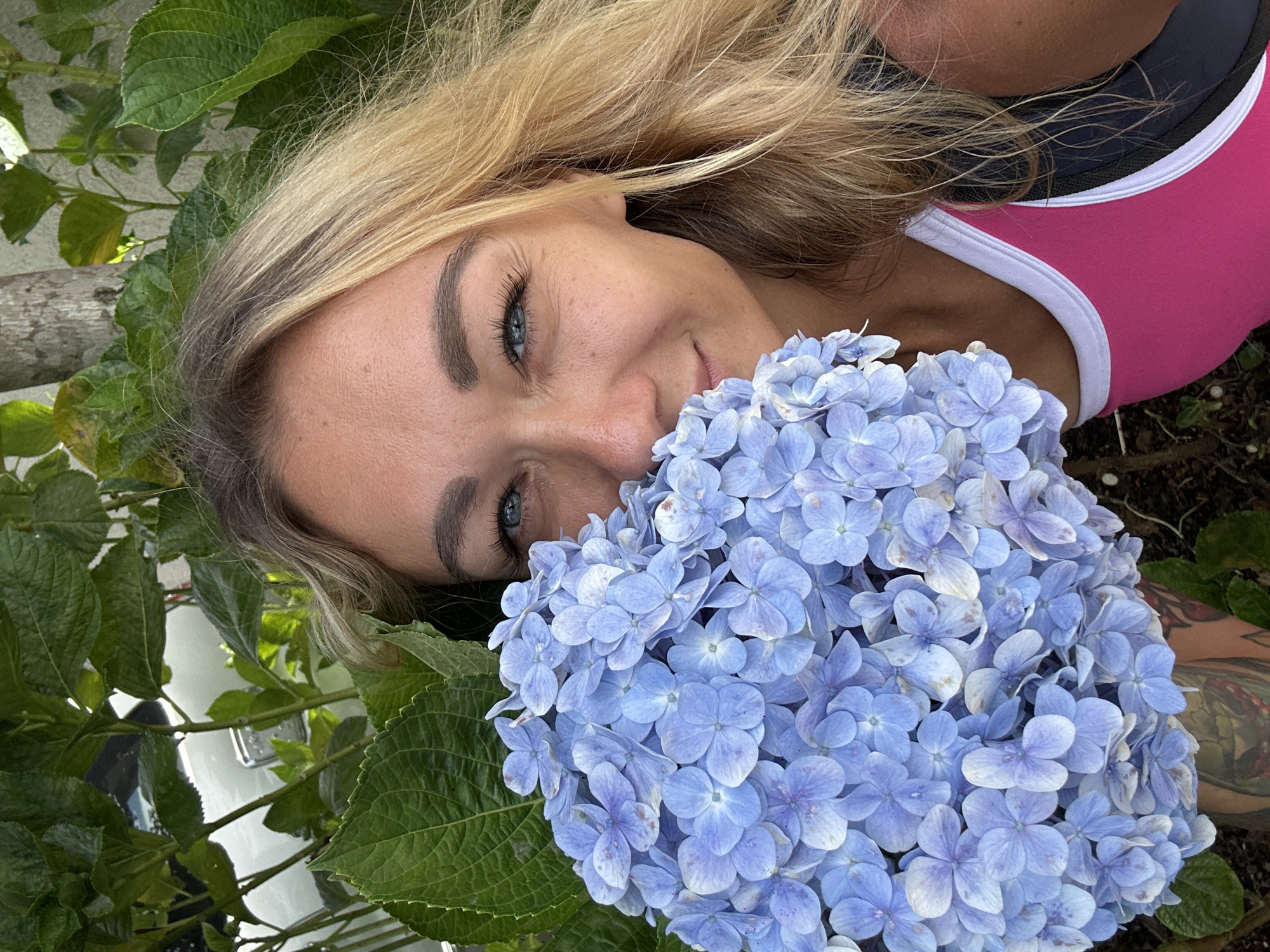 A blonde woman is holding a large bunch of light blue hydrangeas. She is smiling and looking towards the camera. The woman is wearing a pink and white tank top with a black sports bra underneath. She has a tattoo visible on her arm. The background consists of green leaves and a light-colored wall. The image is taken outdoors, possibly in a garden or park setting. There is no specific indicator of the city of Da Lat, Vietnam.
