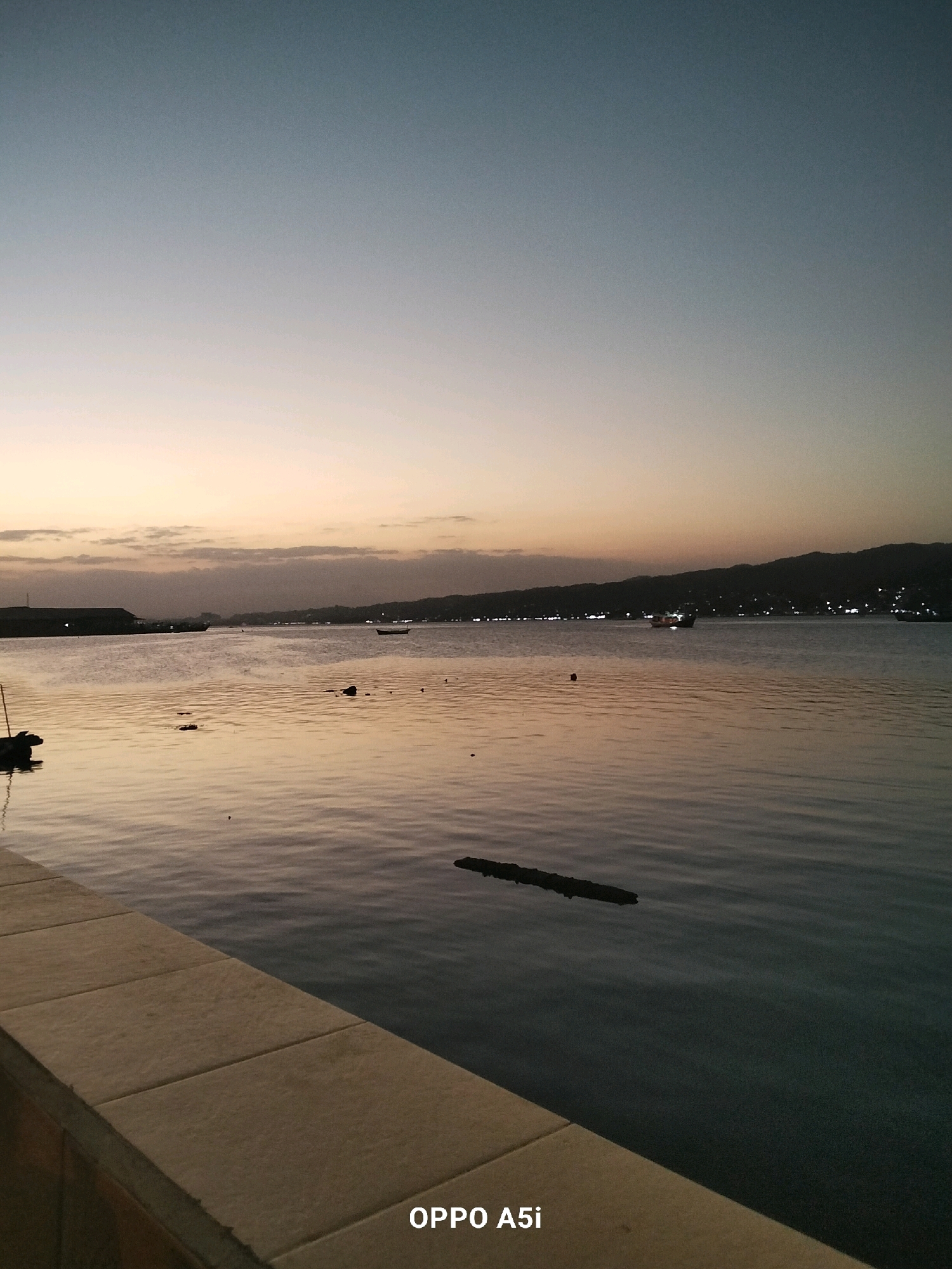 The image captures a serene waterfront scene at dusk in Kendari, Indonesia. The sky exhibits a gradient of soft blues and oranges, transitioning into a dusky hue. The calm water reflects the subtle colors of the sky and the distant lights of the city along the coastline. Several boats are visible on the water, some appearing to be moored while others are in motion. The foreground features a stone embankment, suggesting a public promenade or harbor area. The overall atmosphere is peaceful and tranquil, characteristic of an evening by the water.