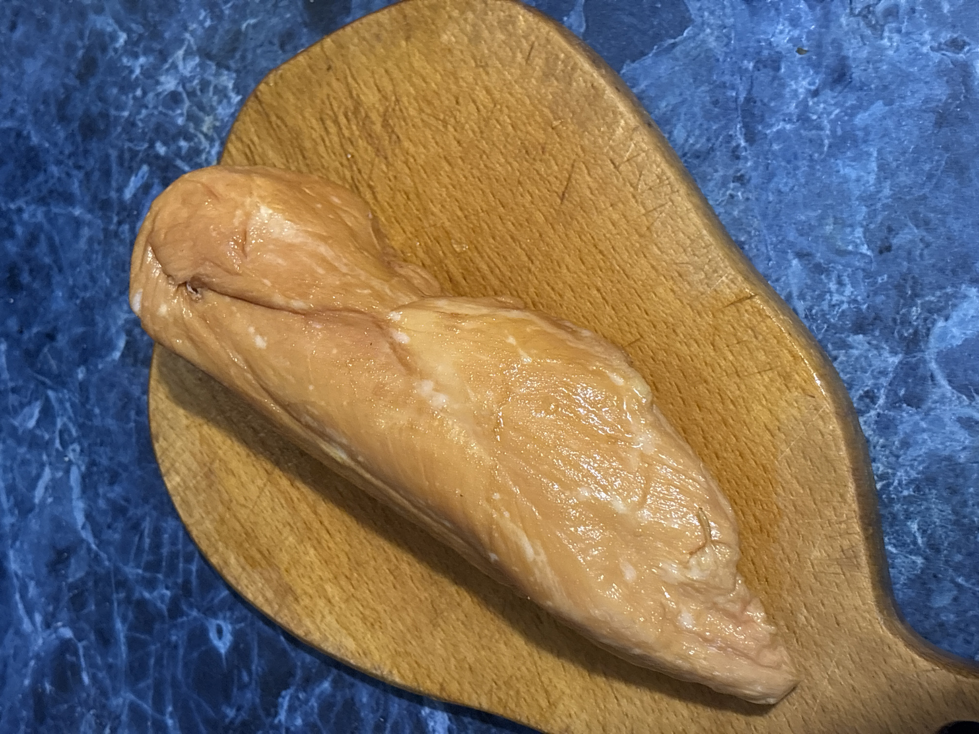 A single, raw chicken breast lies on a wooden cutting board. The cutting board is oval-shaped with a handle. The background is a blue, marbled countertop. The image is taken from a slightly elevated, overhead angle. The chicken breast is elongated and appears to have a light, translucent color with some white speckles. The cutting board has visible knife marks and a worn appearance. The blue countertop has a subtle marble pattern with lighter blue and white veins. The lighting suggests an indoor setting with overhead light source casting subtle shadows. No people or specific locations within Kremenchuk, Ukraine are identifiable in this image.