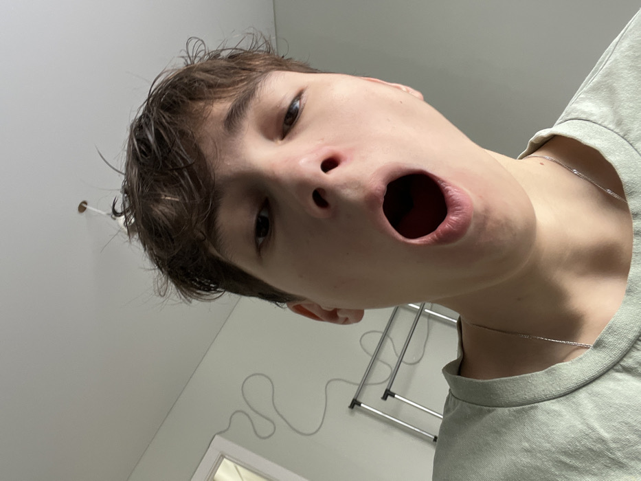 The scene depicts an interior room in Zainsk, Russia. A young individual, of indeterminate gender, with fair skin and disheveled dark hair, is positioned centrally in a low-angle photograph, looking directly into the camera. The individual's mouth is wide open, revealing the oral cavity. They are wearing a light green crew-neck t-shirt and a thin silver-colored chain necklace. The background consists of plain, light-colored walls and a white ceiling. On the left wall, a white electrical cable is affixed in an undulating pattern. Below the cable, a partial view of a multi-tiered tubular metal rack is visible. A doorframe is present in the upper left, and a ceiling-mounted light fixture base with exposed wiring is visible near the top center of the frame.
