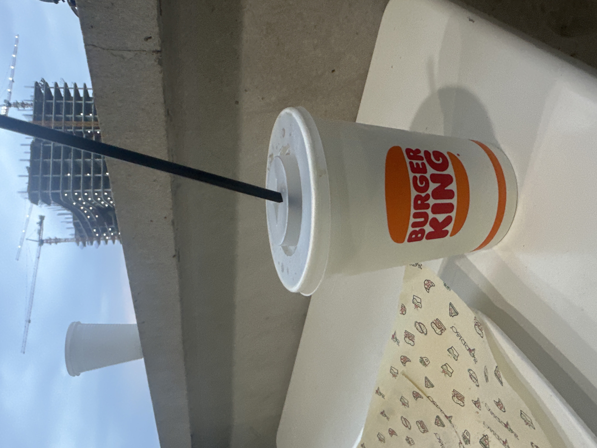 The scene is a Burger King restaurant in an unknown city. A Burger King beverage cup with a straw is on the table. Outside the window is a construction site of a skyscraper. There are construction cranes visible.