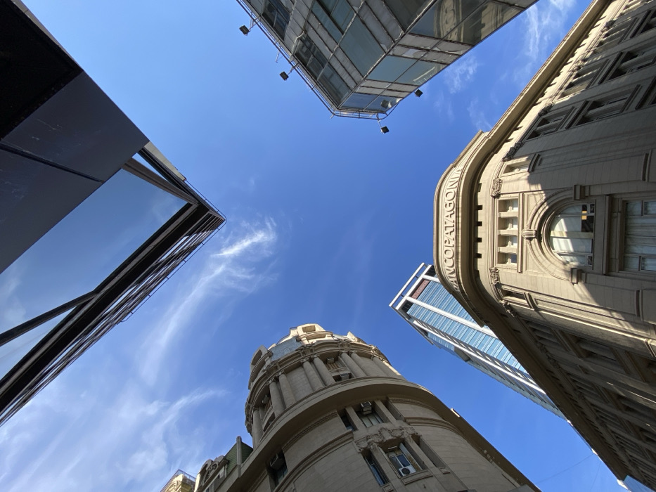 A low-angle shot captures several buildings against a bright blue sky with wispy clouds. The buildings range in architectural style from classical to modern, including one labeled "BANCO PATAGONIA." The composition creates a frame-like effect, drawing the eye towards the sky above. The sunlight illuminates different facades.