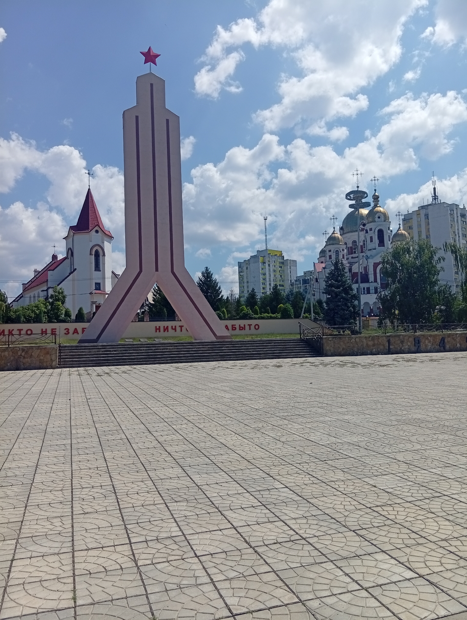 The image shows a sunny day in Rybnitsa, Moldova. In the foreground is a large, paved plaza with a repeating geometric pattern. In the mid-ground, a large, light-colored monument with a red star on top stands prominently. To the left of the monument is a white church with a red roof and a cross. To the right of the monument, a large building with golden domes and crosses, resembling a cathedral, is visible. In the background, there are several multi-story residential buildings and trees. Red lettering is visible on a wall behind the monument, which appears to read "НИКТО НЕ ЗАБЫТ" (Nobody is forgotten) and "НИЧТО НЕ ЗАБЫТО" (Nothing is forgotten). The sky is blue with scattered white clouds.