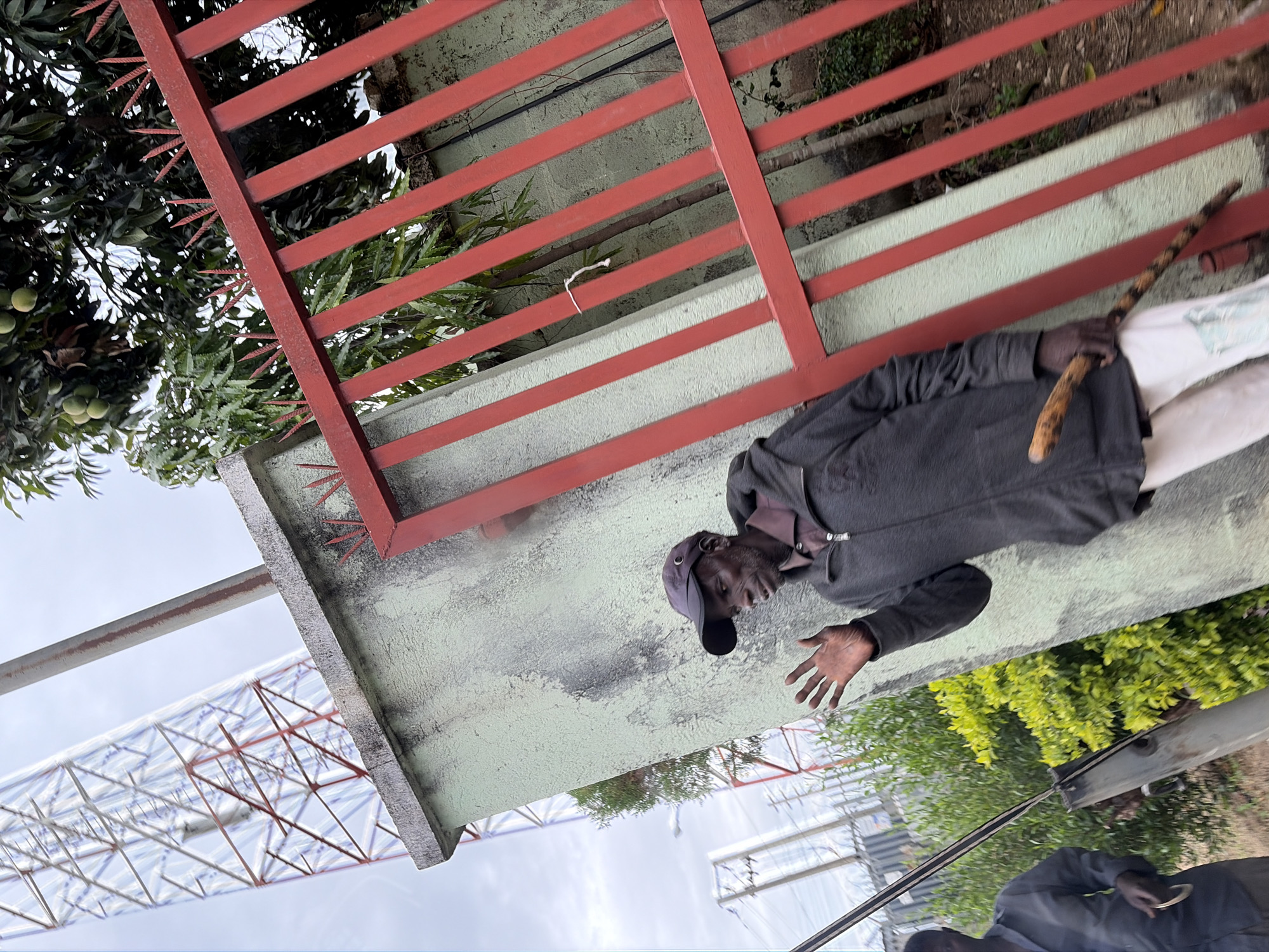 A man wearing a dark grey jacket, dark pants, and a dark cap is standing in front of a green concrete wall with a red metal fence on top. He is holding a wooden stick in his right hand and extending his left hand outwards. Behind the wall, there is a mango tree with green mangoes visible. In the background, there is a white and red metal structure, possibly a tower. The image was taken at an angle, making the ground appear to be the wall. The location is Gura Top, Nigeria.
