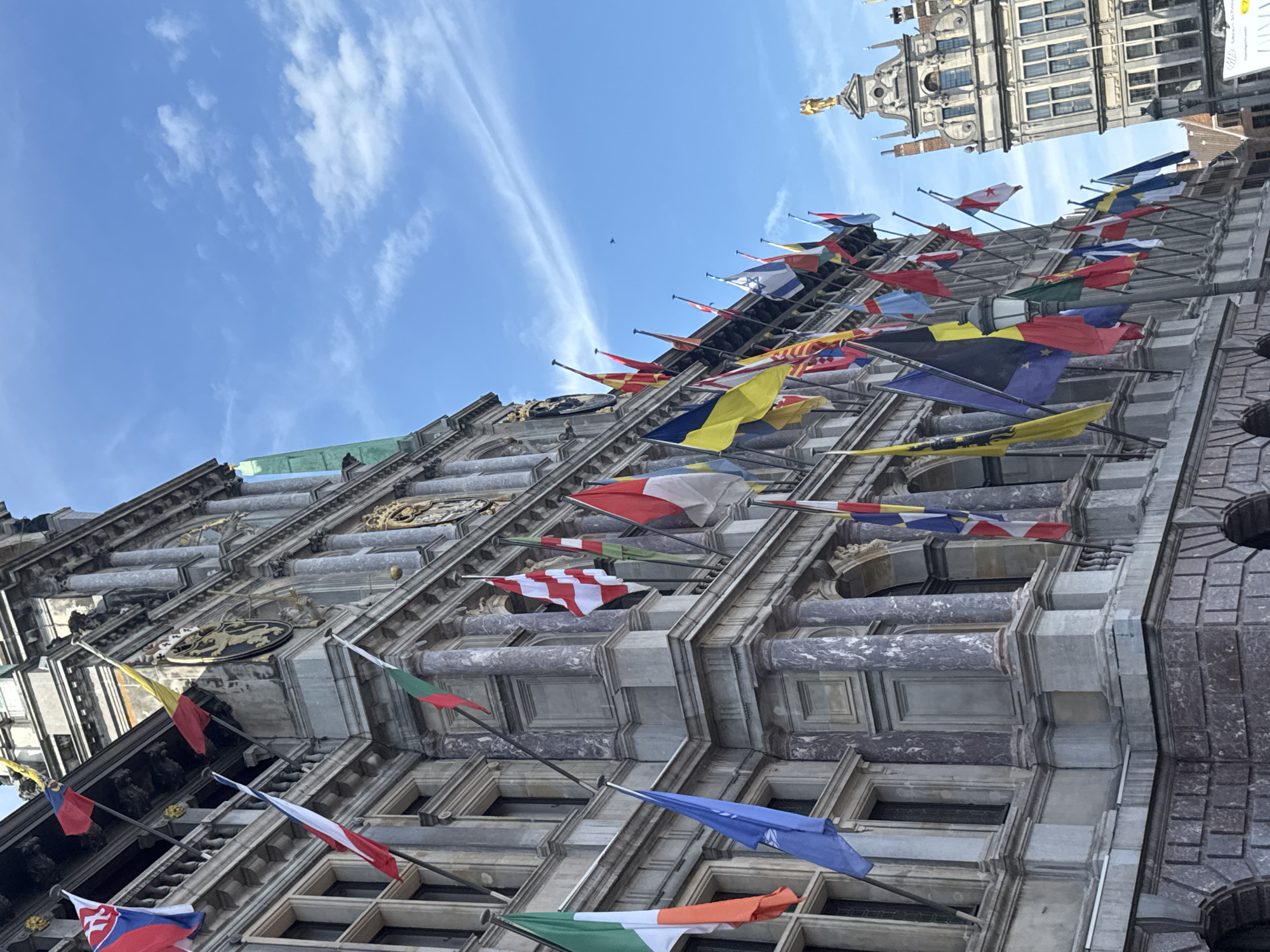 The image depicts a low-angle view of the Antwerp City Hall, a historic building located in Antwerp, Belgium, adorned with numerous national flags displayed on poles extending from its facade. The architecture of the building is ornate, with columns, detailed stonework, and multiple stories. The sky is bright blue with scattered white clouds. In the background, another building with a golden statue on top is visible. The flags represent various countries, including what appears to be the Ukrainian flag (blue and yellow) and the Belgian flag (black, yellow, and red). The overall scene suggests a formal occasion or celebration, with the flags prominently featured against the backdrop of the city's architecture. There are no people visible in the image.