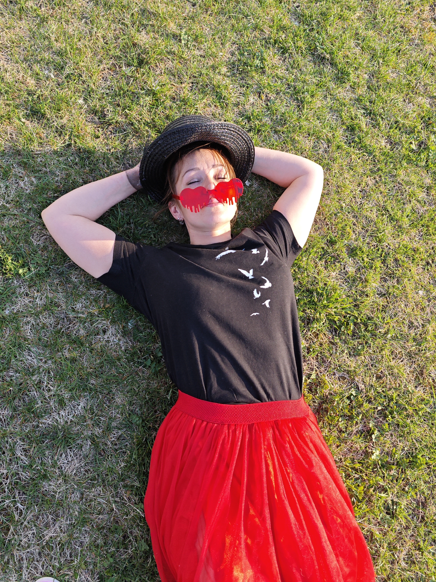 A young adult woman is lying on her back in a grassy area, presumably in Markovo, Russia, given the prompt. She has her arms raised and crossed behind her head. The woman is wearing a black t-shirt with a flock of white birds printed on it and a long red skirt. She is also wearing a black hat and red heart-shaped glasses with dripping paint. The grassy area appears to be somewhat patchy with both green and brown grass.